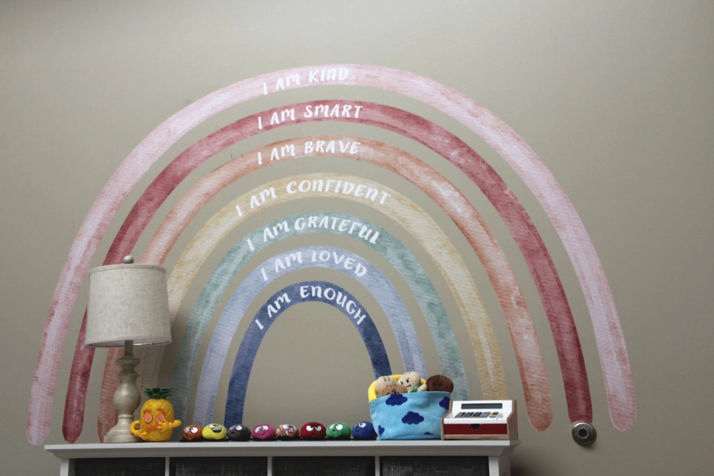 Decorative wall with a rainbow mural containing positive affirmations, including 'I am kind,' 'I am smart,' 'I am brave,' 'I am confident,' 'I am grateful,' 'I am loved,' and 'I am enough.' There is a table below with plush toys, a toy cash register,