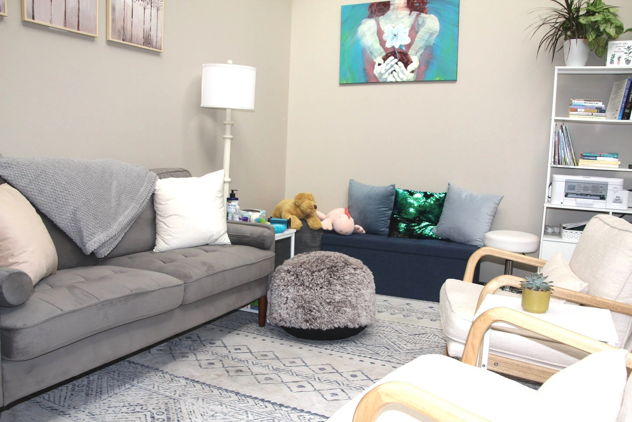 Therapy room with gray and navy furniture, a round fuzzy ottoman, a white bookshelf, and a wall art with a floral theme.