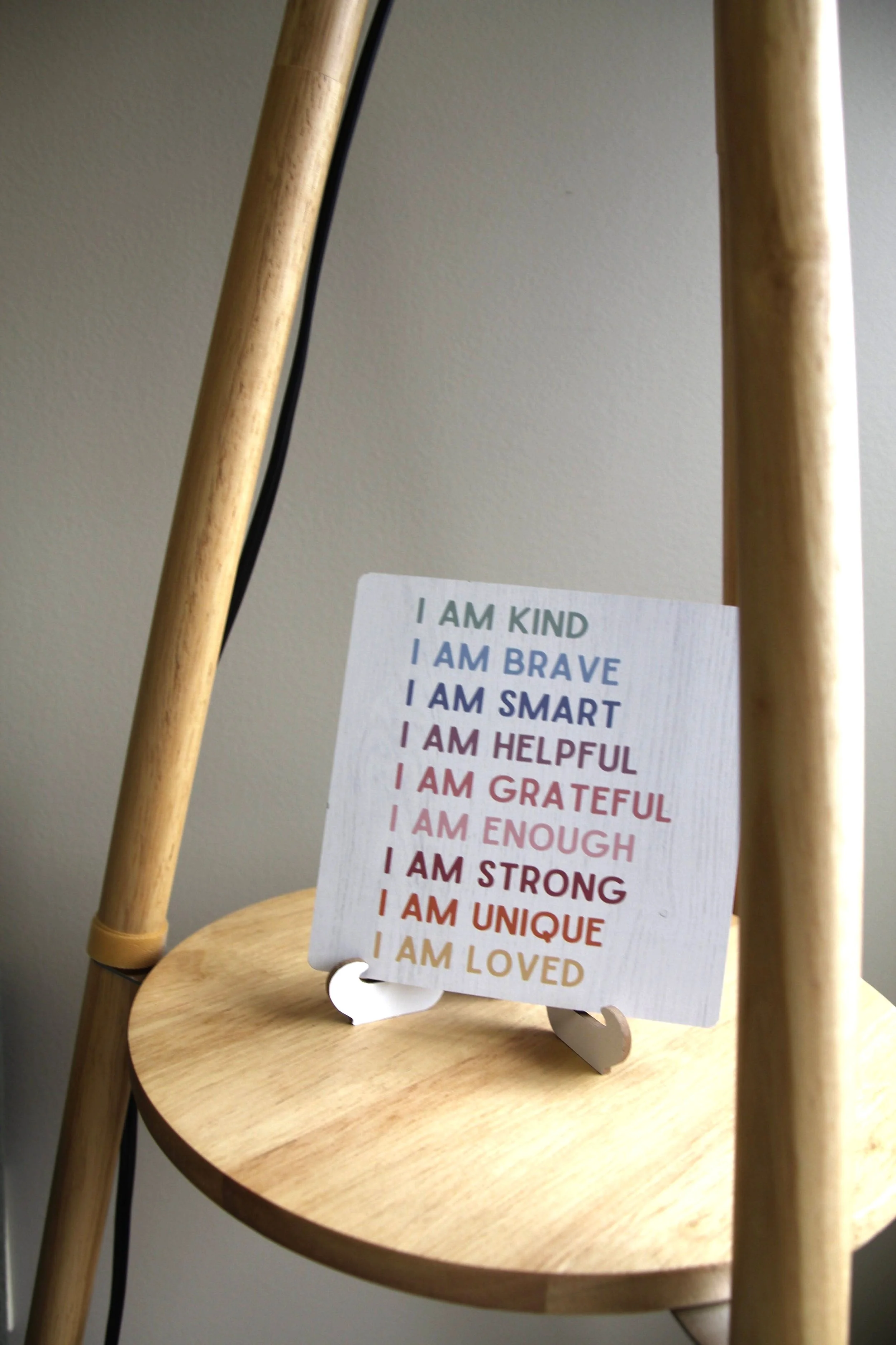 A small sign with positive affirmations on a wooden stand, placed on a round wooden surface, with parts of a wooden ladder or furniture in the background.