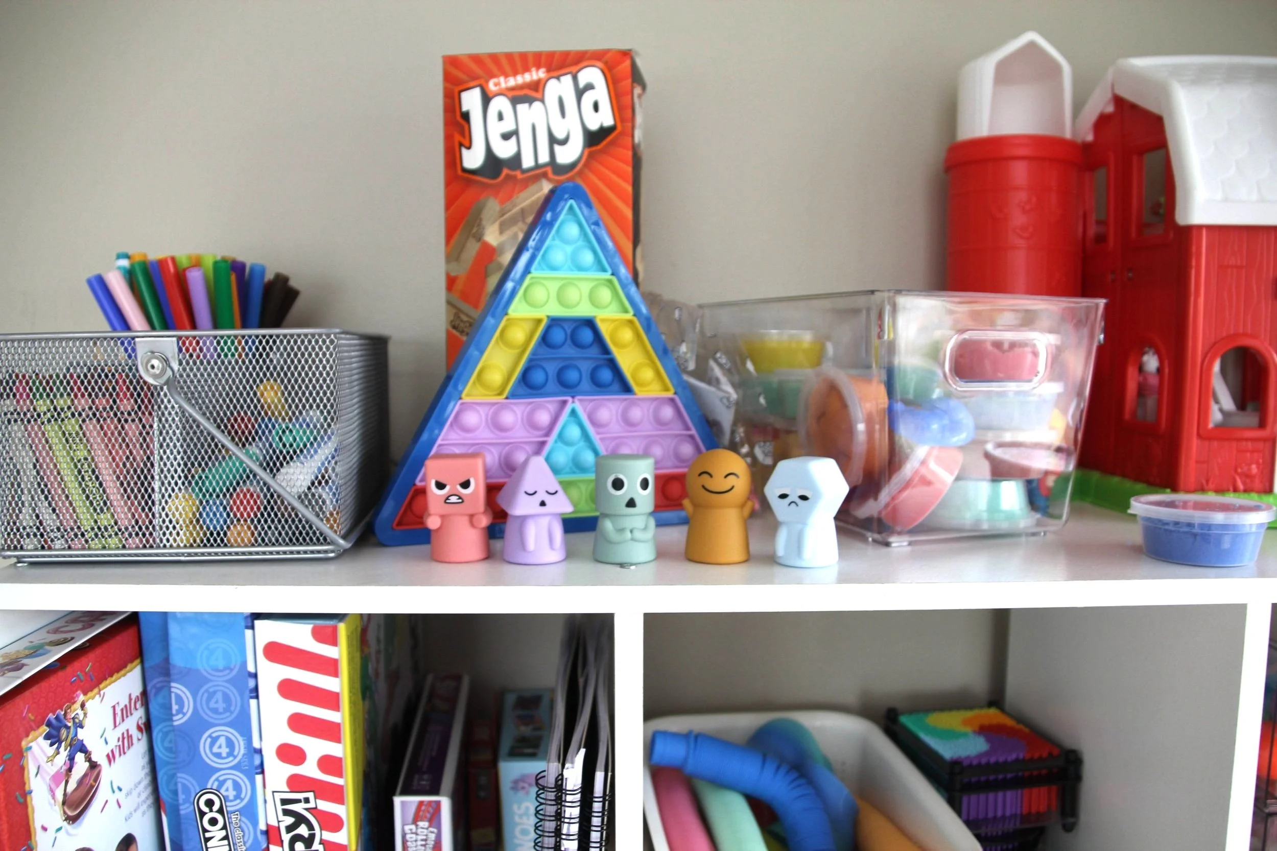 A shelf filled with various toys and games, including a Jenga game, colorful marker pens, a set of foam puzzle pieces, and small figurines resembling characters. There are also storage containers with other toys and arts and crafts supplies.