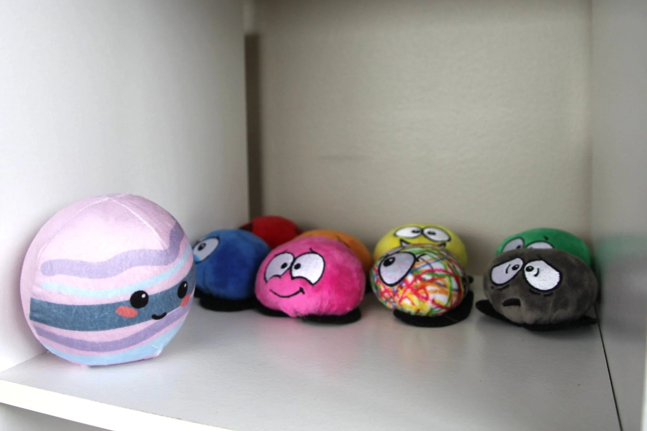 A collection of colorful, plush, and cartoon-like rocks with expressive faces on a white shelf.