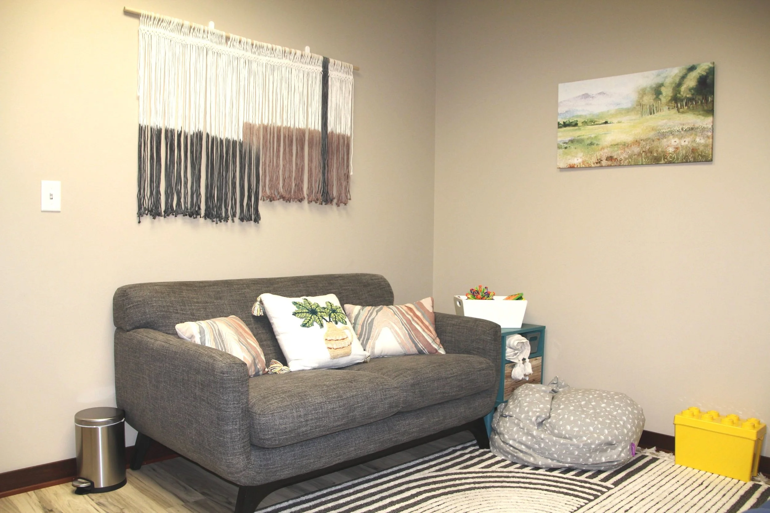 Therapy office with gray sofa, decorative pillows, a small side table with a white container, a bean bag chair, a yellow plastic storage bin, and a striped rug. There is a window with a multi-colored fringed curtain, a landscape painting on the wall