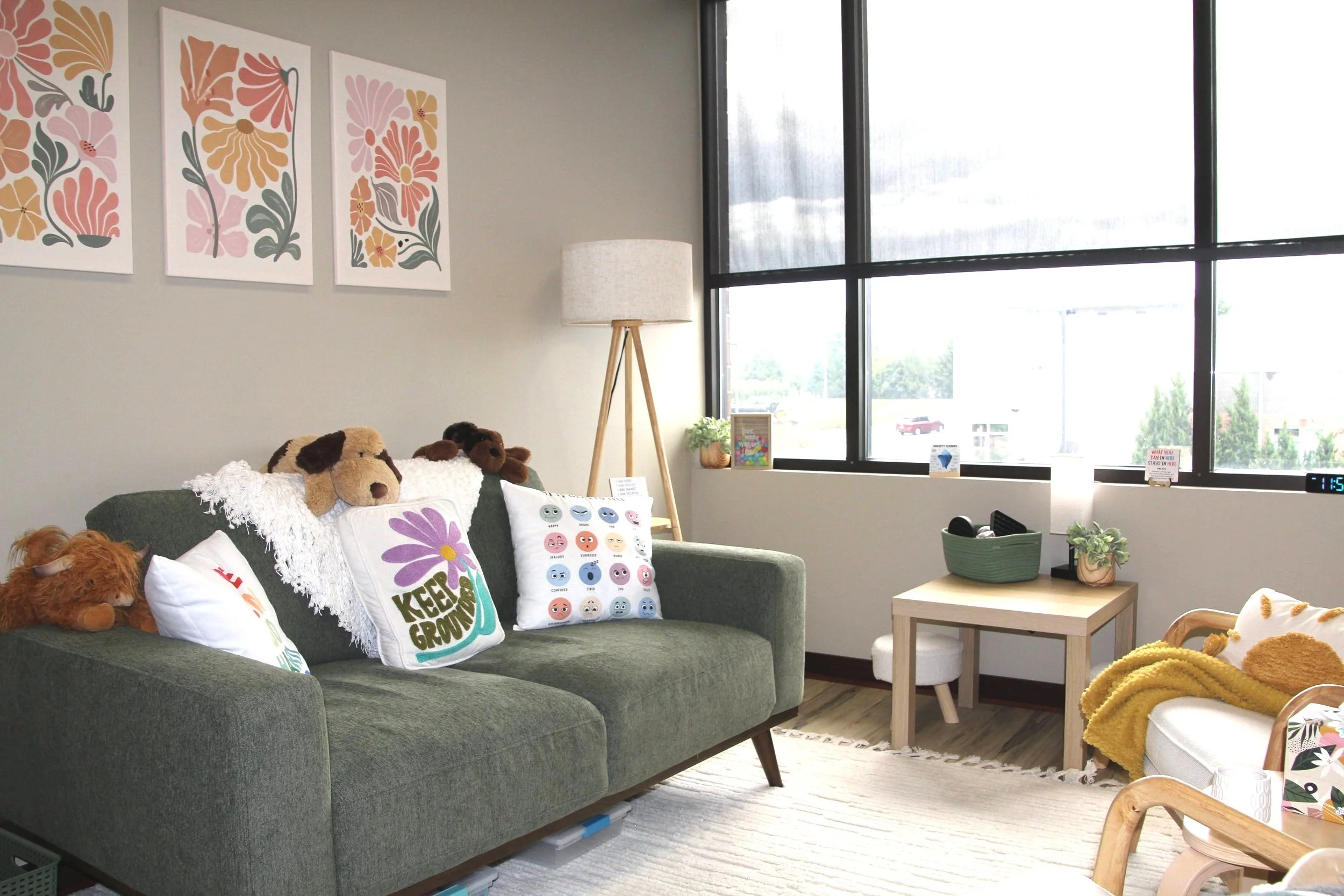 Cozy therapy office with a dark green sofa, decorated with plush animal pillows and a white pillow with colorful designs, a light wooden side table, large windows with black frames, and wall art featuring floral patterns in pastel colors.