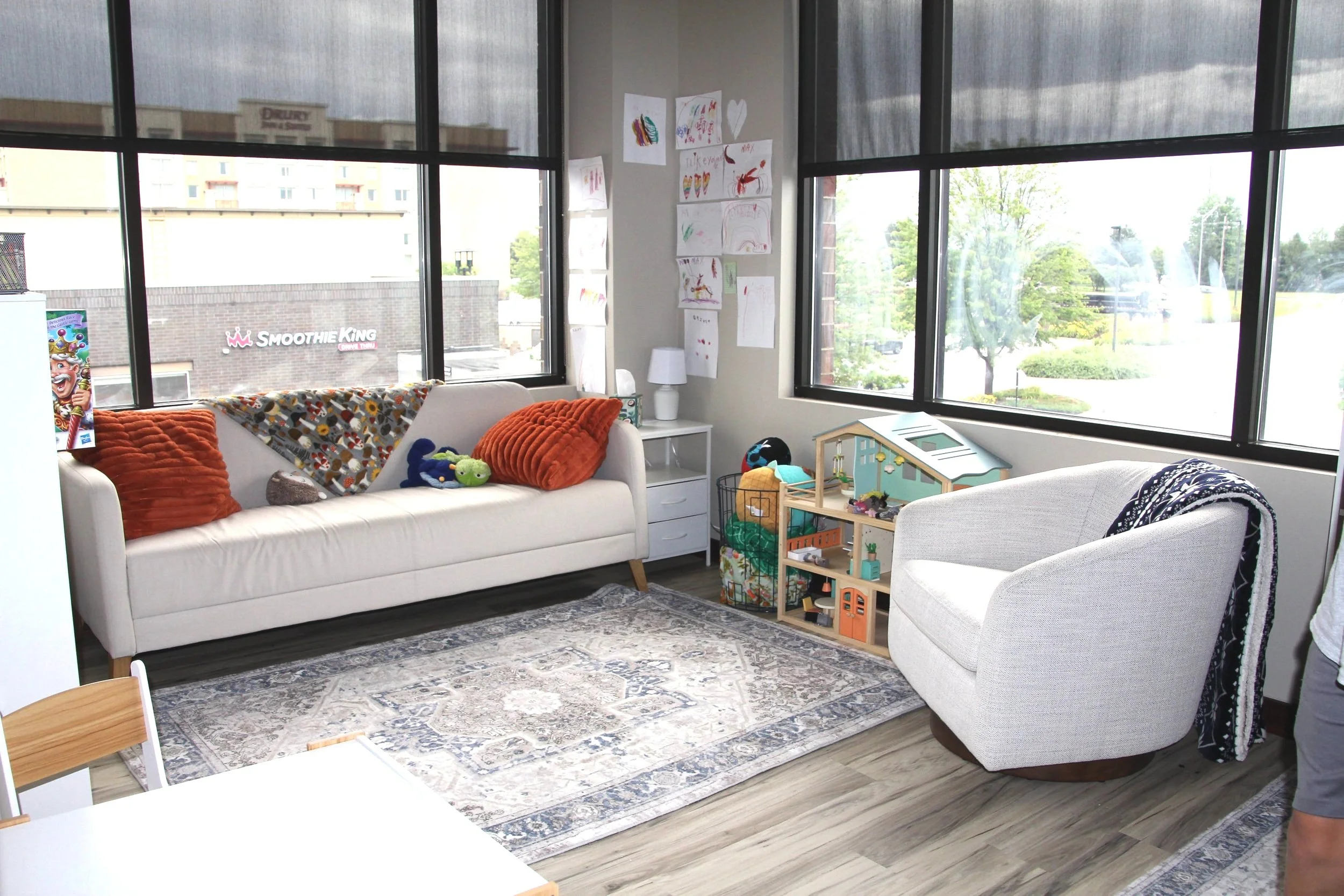 Bright therapy room with large windows, white sofas with orange and gray pillows, children's toys, and artwork on the wall.