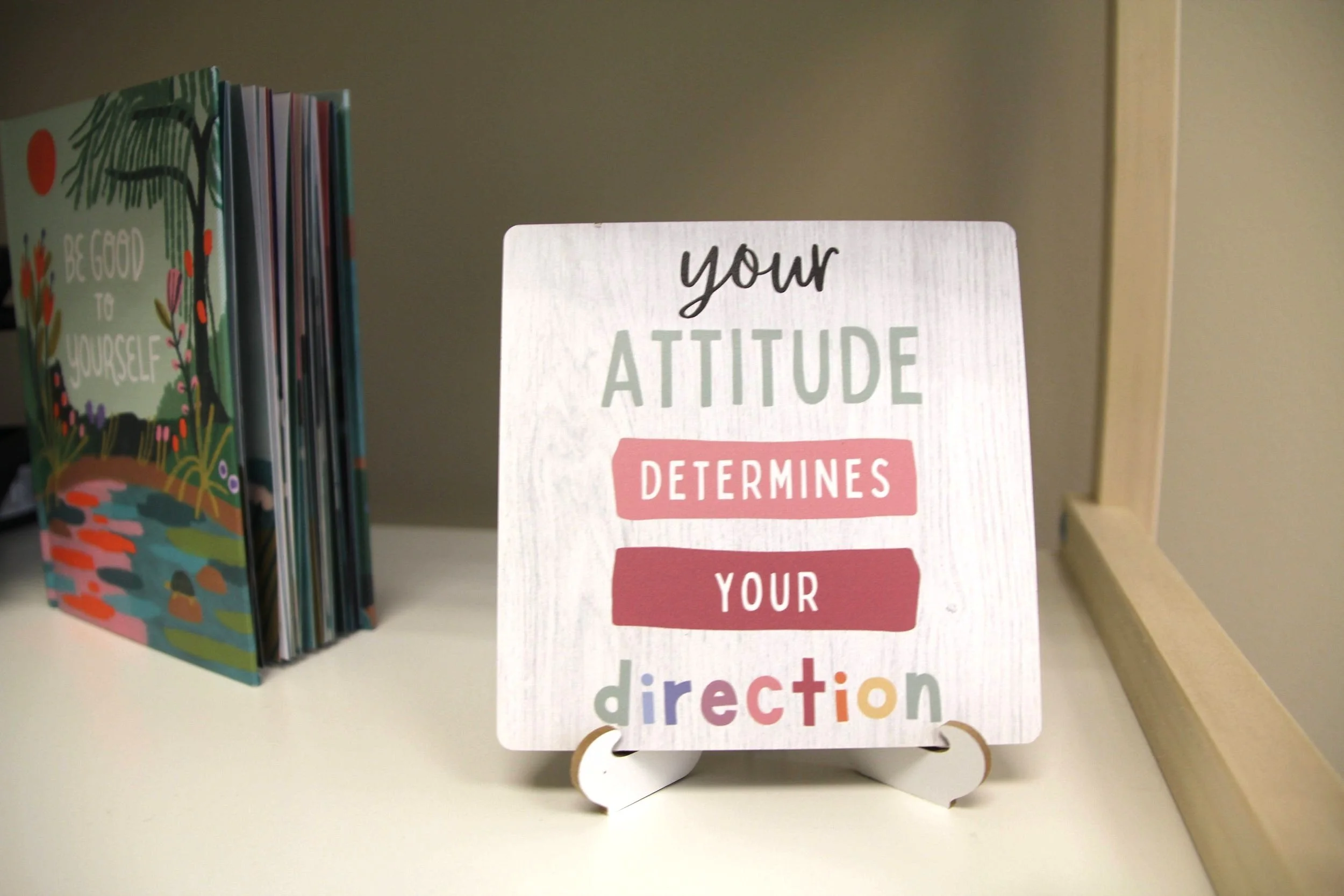 A decorative sign reads 'your attitude determines your direction' set on a small white stand on a white surface, with a colorful book featuring nature illustrations nearby.