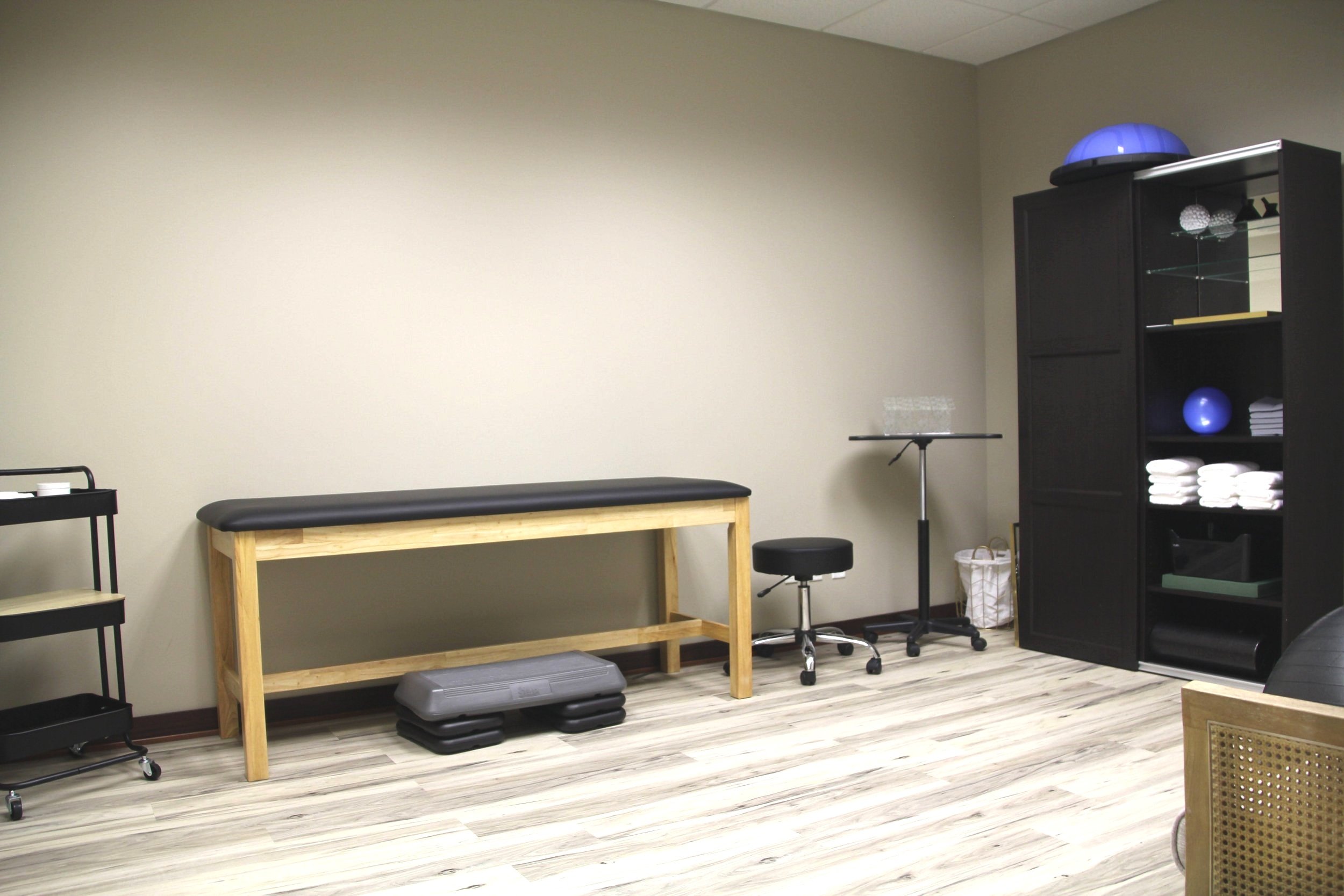 Therapy or massage room with a black massage table, a step platform, a rolling stool, a black cabinet filled with towels and therapy equipment, and a small table with water glasses in a plain room with light-colored walls and wood-patterned flooring.
