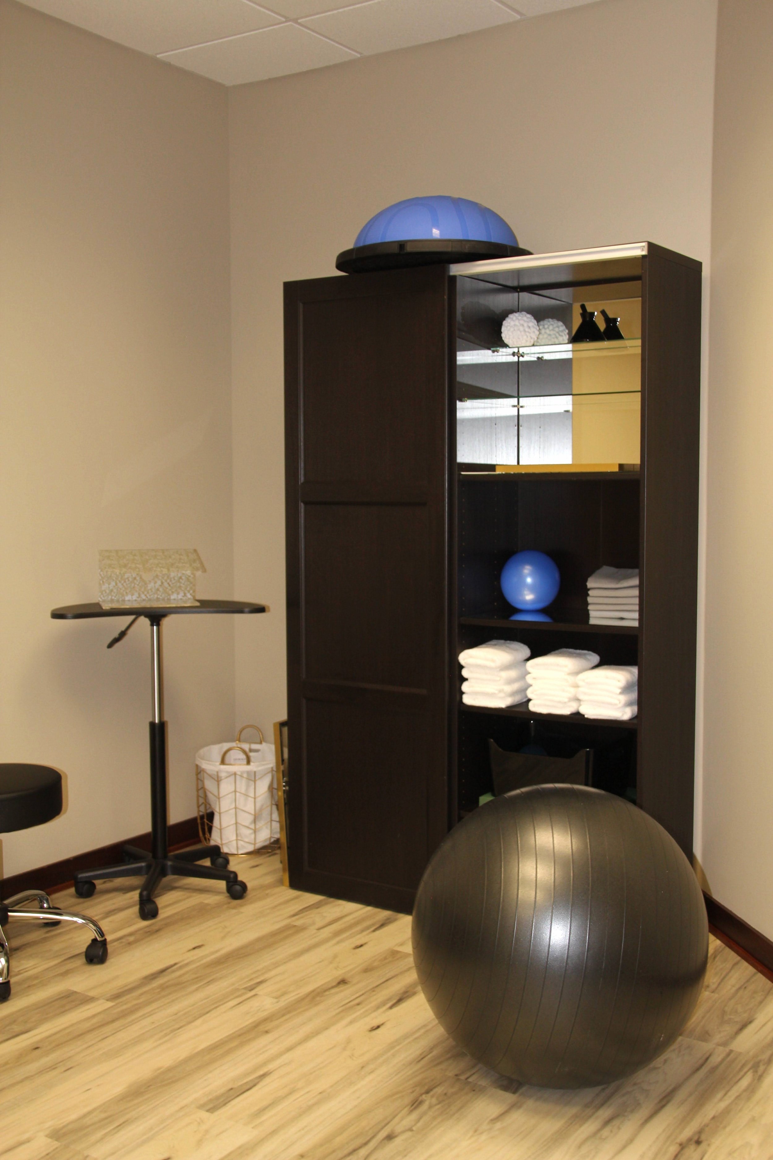 Physical Therapy room with a large black exercise ball on the floor, a dark wooden shelf with folded white towels, a blue exercise ball, and a blue half dome balance trainer on top of the shelf. There is also a small white basket, a black stool.