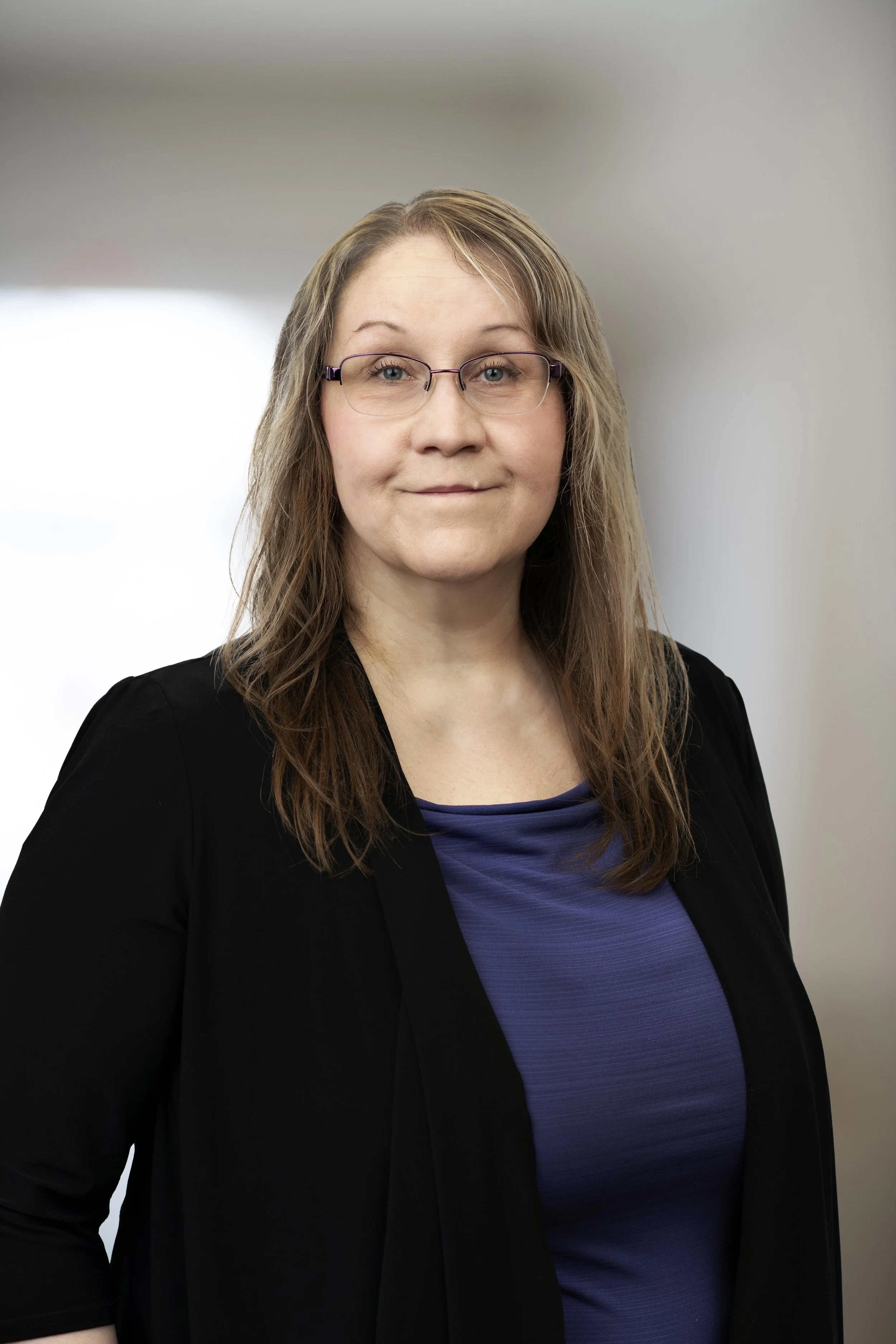 A woman with shoulder-length light brown hair, glasses, wearing a black blazer over a blue top, standing against a blurred neutral background.
