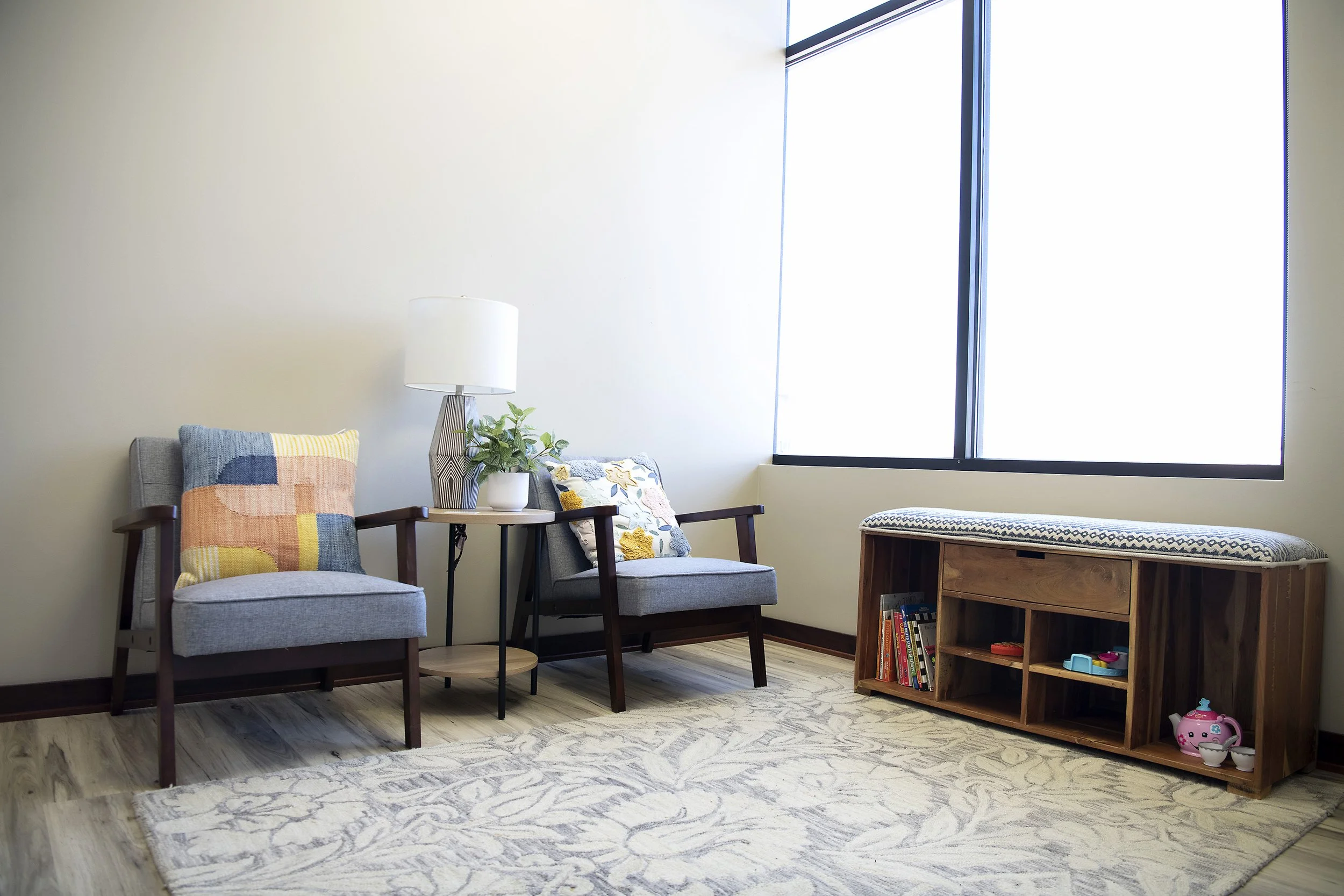 small waiting room with two gray armchairs with colorful pillows, a small round side table. On the table, a white lamp and green plant. Large window with blinds, wooden storage unit with toys, books, and a cushioned top.