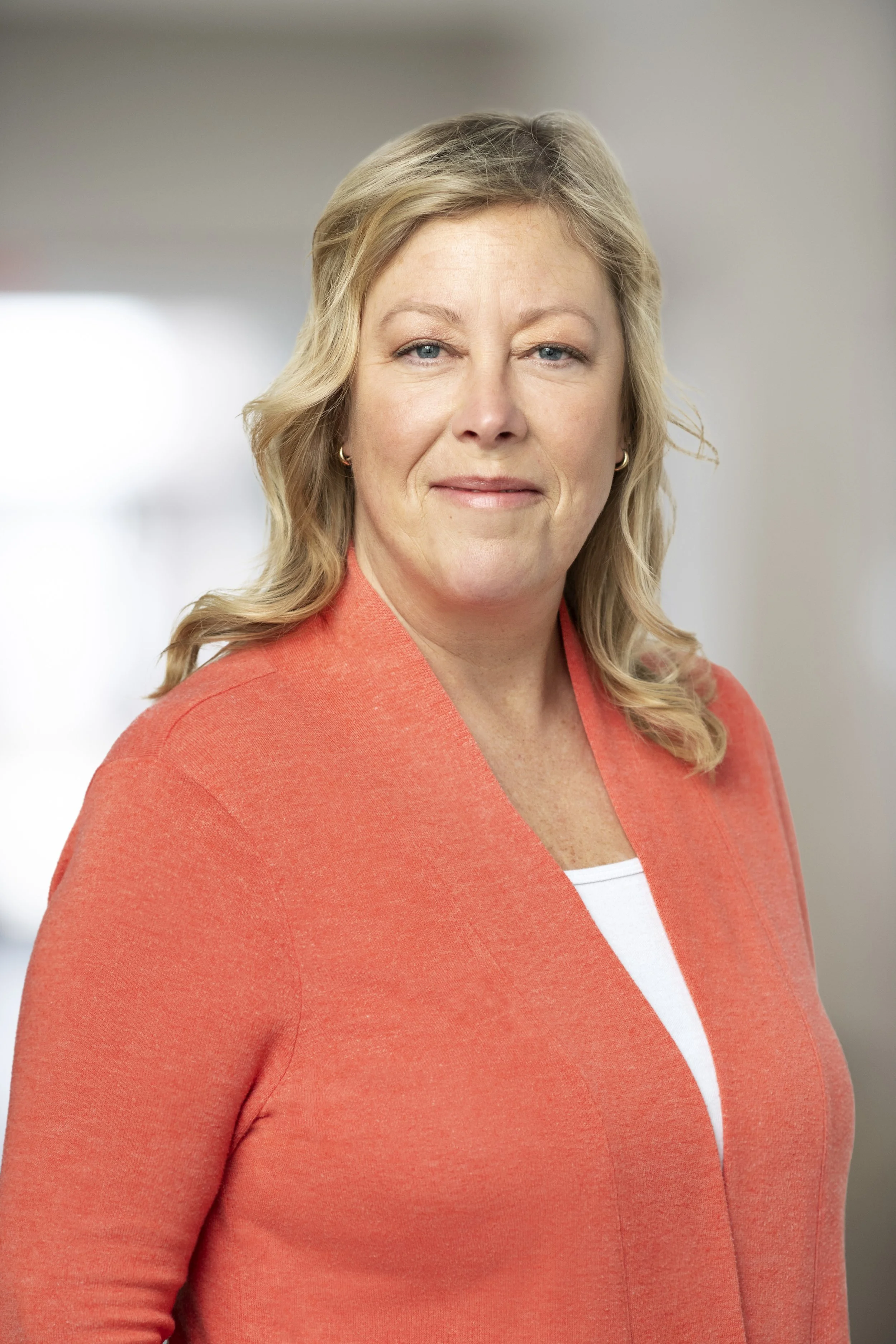 Portrait of a woman with blonde hair wearing a coral blazer and a white top, standing in an indoor setting with a blurred background.