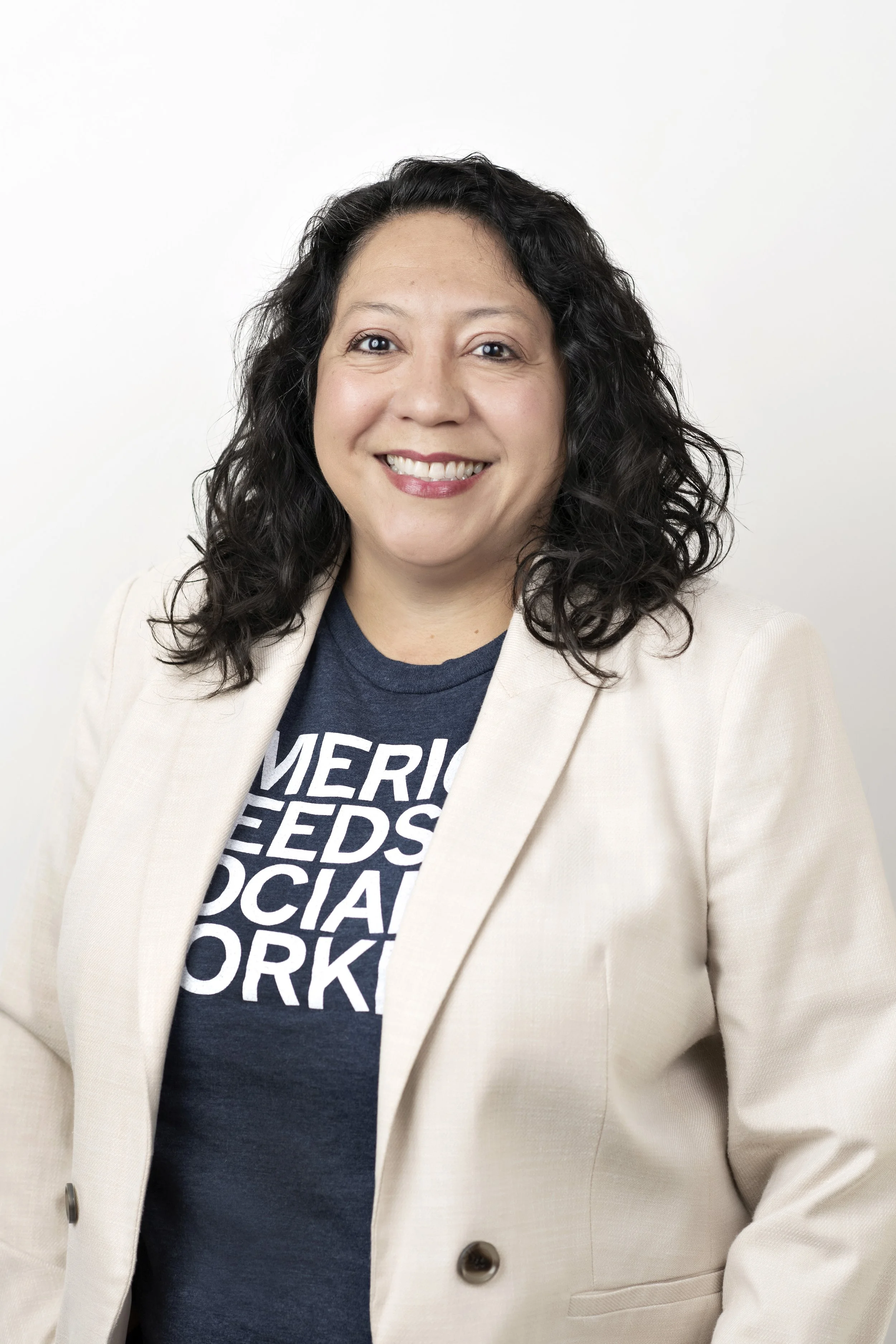 A woman with dark curly hair wearing a white blazer and a navy blue T-shirt with white text, smiling against a plain white background.