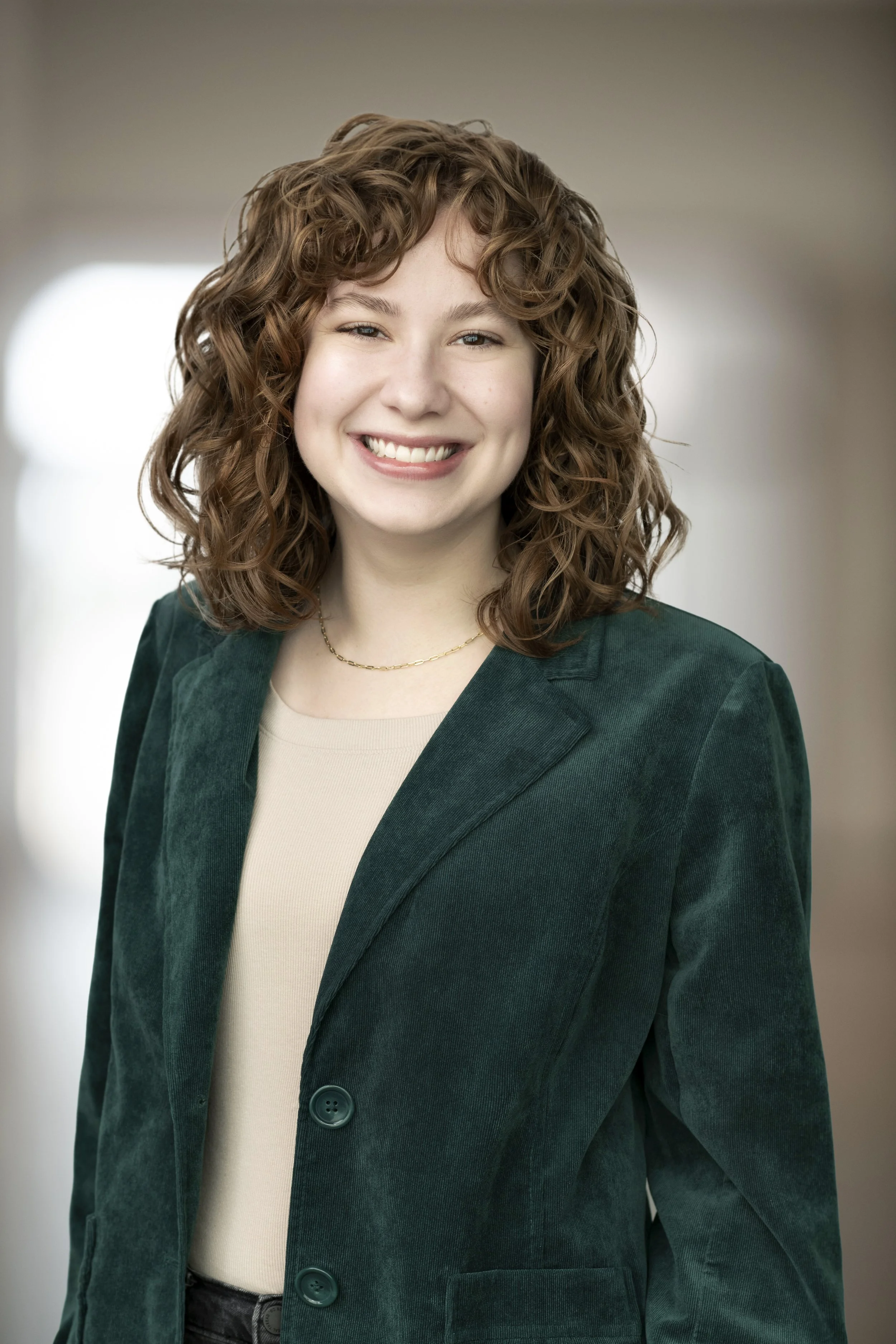 A woman with shoulder-length curly brown hair, smiling, wearing a dark green blazer over a beige top and a gold necklace, in an indoor setting with blurred background.