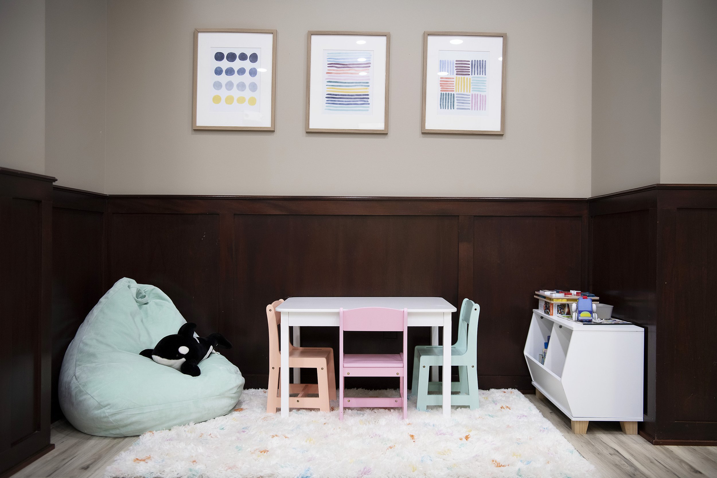 Children's playroom in waiting room with a white table, pastel colored chairs, a white bookshelf with toys and books, a plush bean bag with a stuffed orca, a white fluffy rug, and three colorful abstract art pieces on a beige wall.