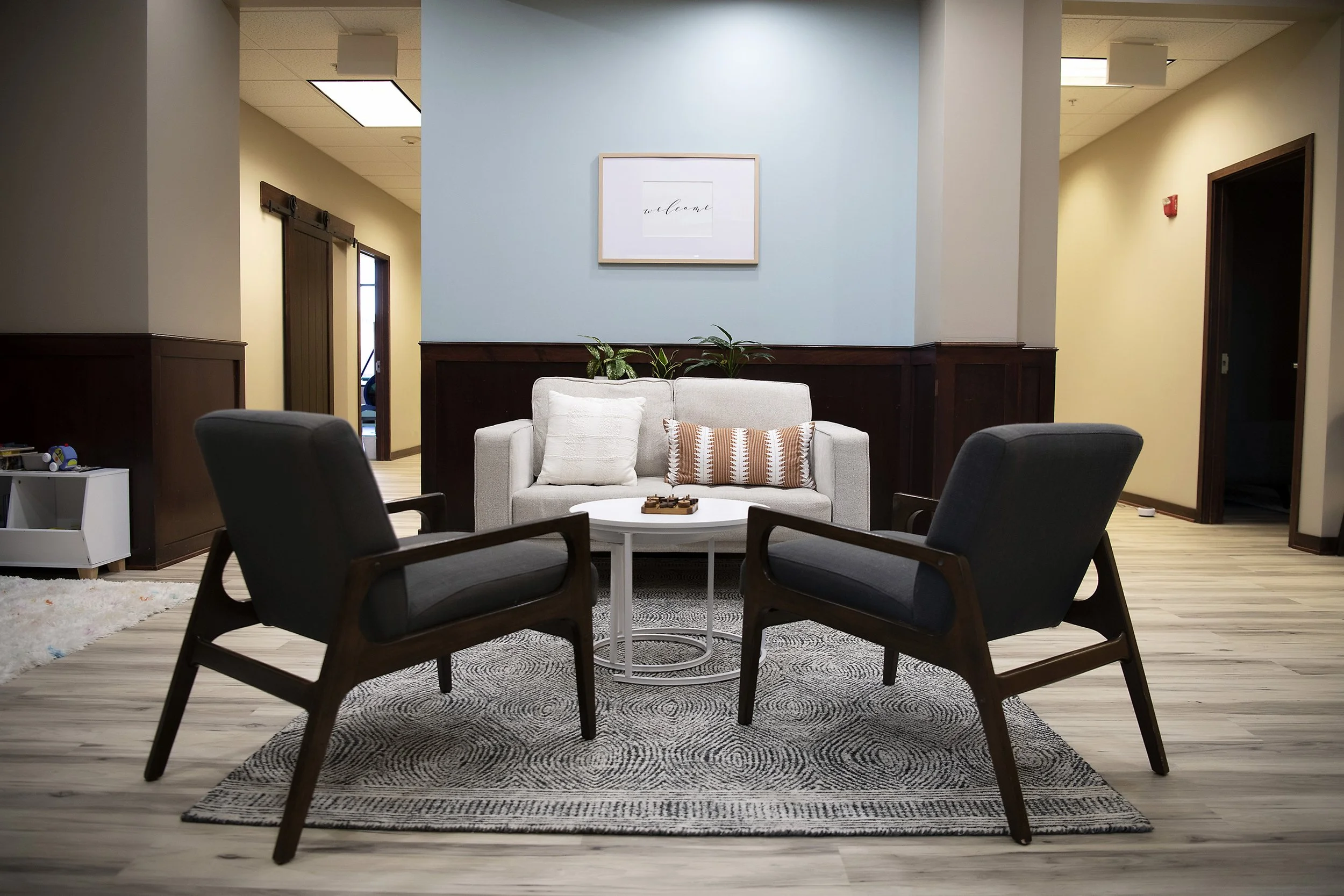 A waiting room with two dark gray armchairs, a white sofa with pillows, a small white round table with a tray, and a patterned area rug on light wood flooring. There are plants and a picture on a light blue wall in the background.