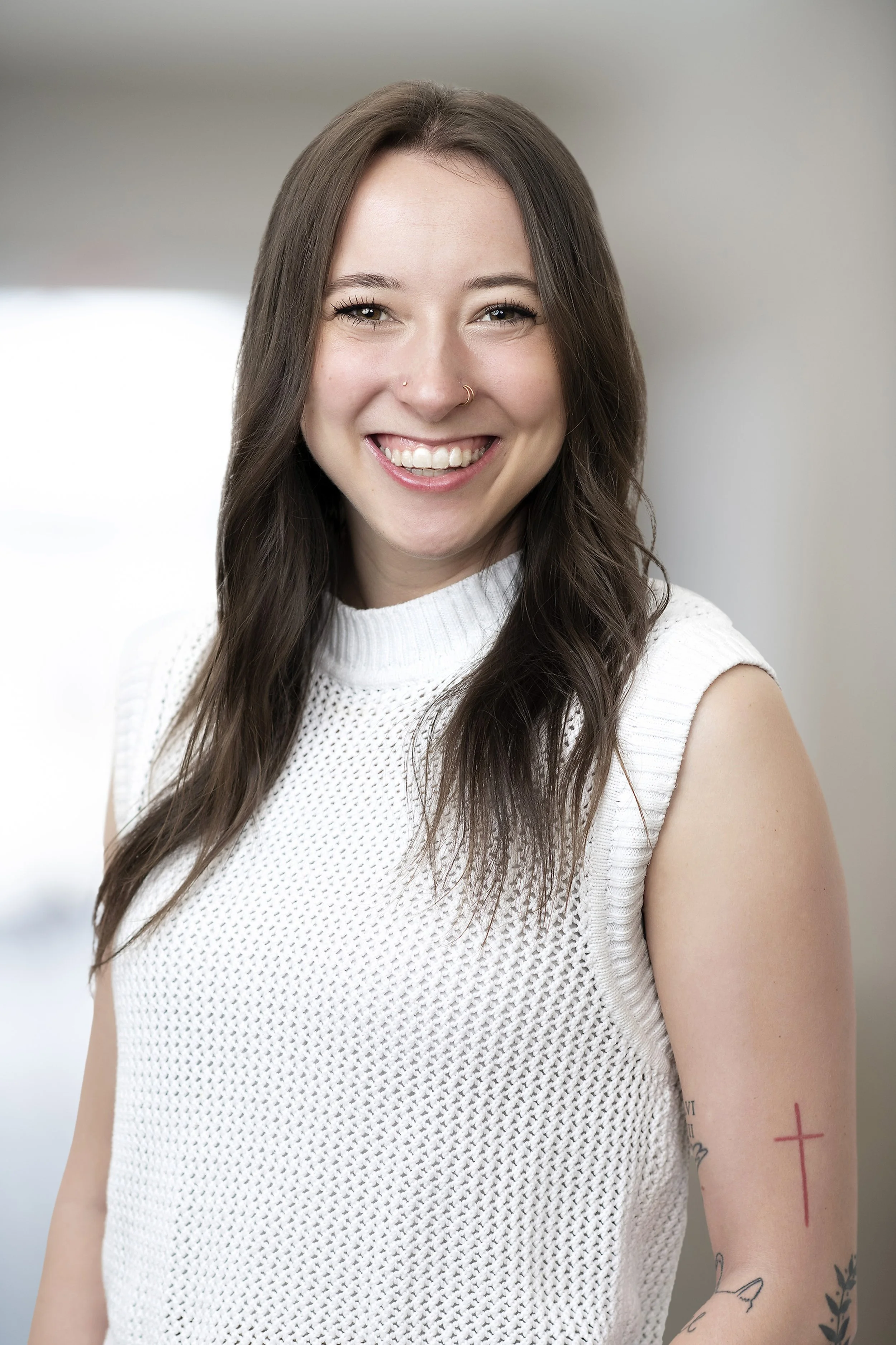 A young woman with long brown hair smiling, wearing a white sleeveless knit top with tattoos visible on her arm, in a bright indoor setting.