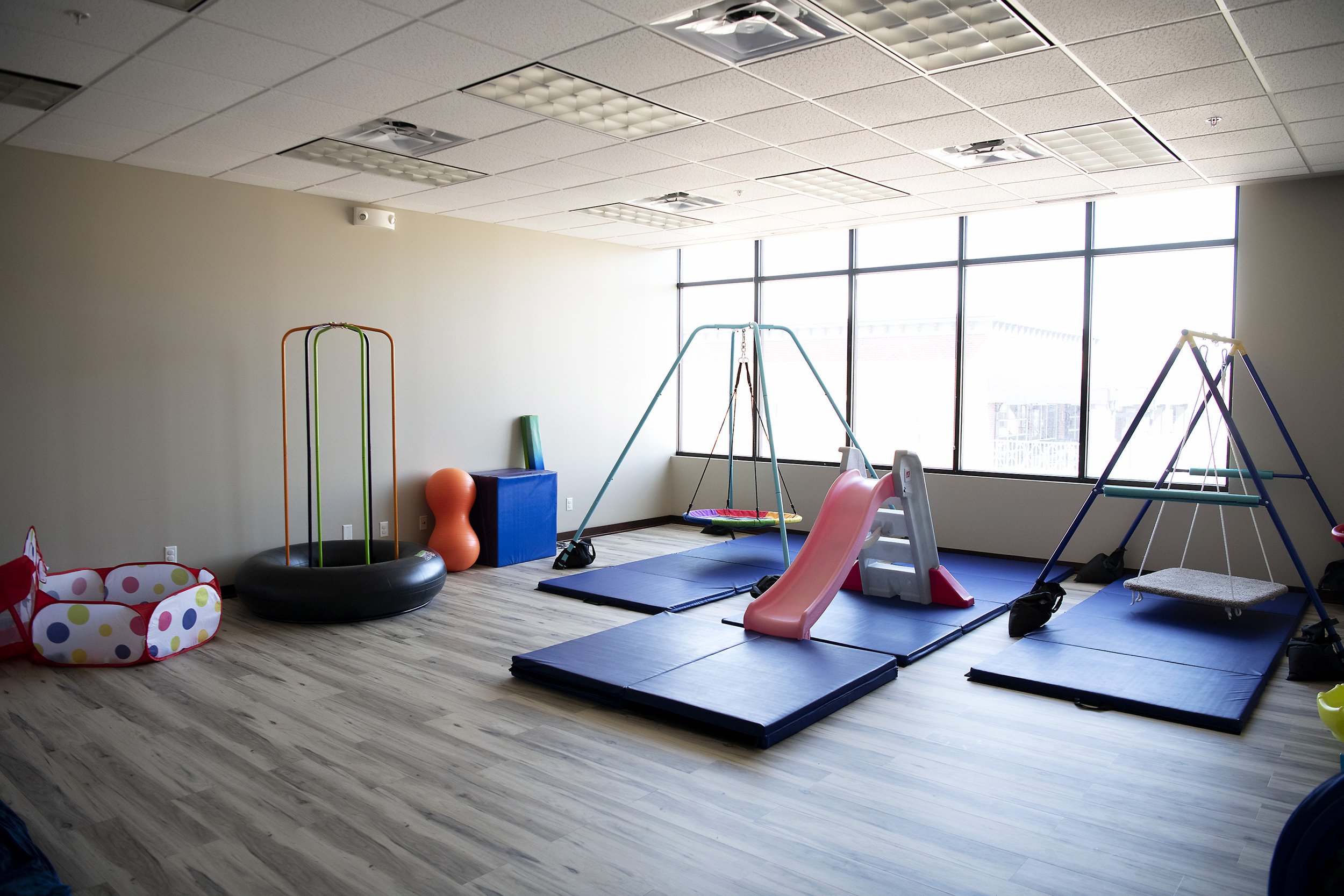 Indoor children's playroom with swings, slide, mats, and toys near large windows for pediatric occupational, speech and physical therapy.