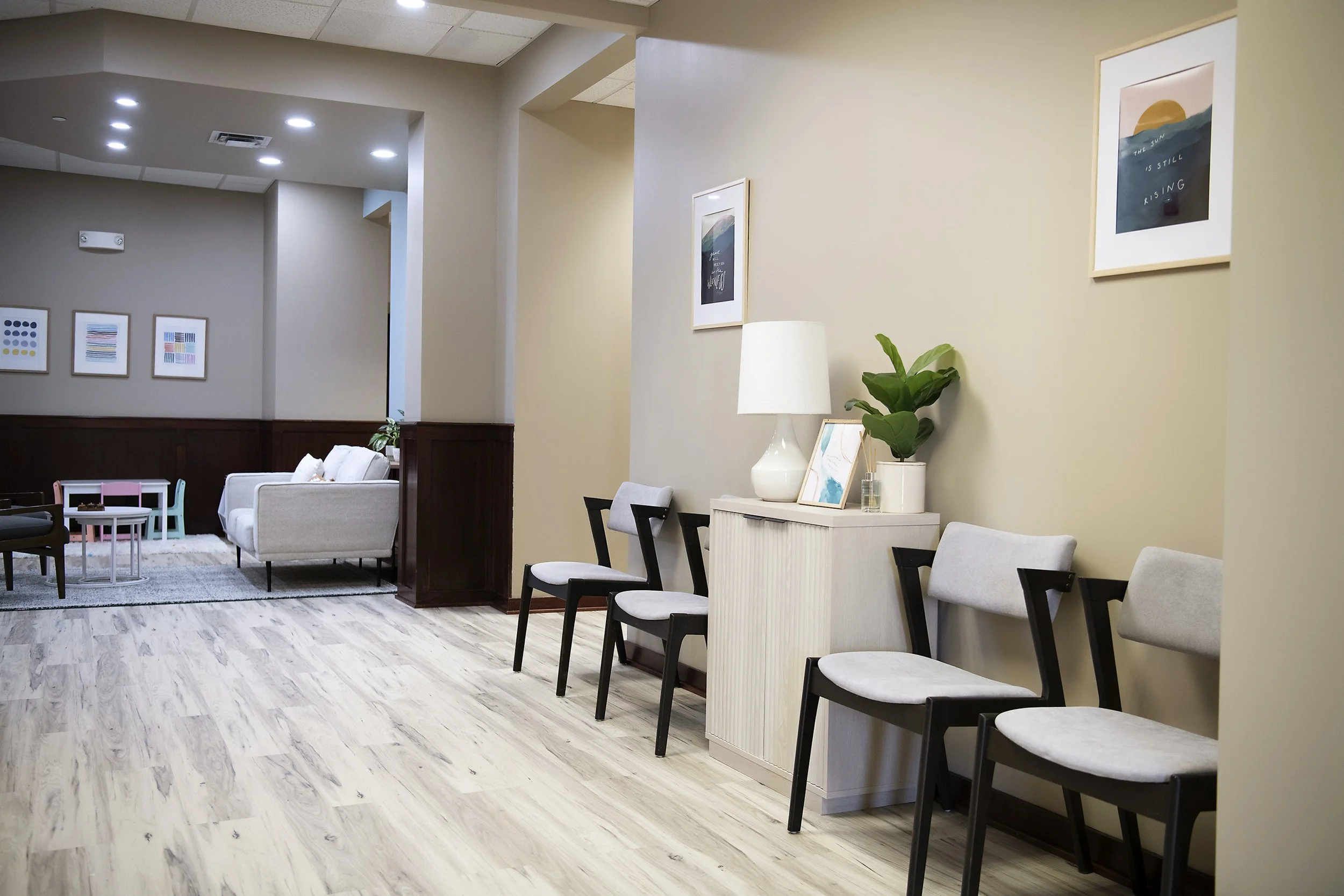 A waiting room with four chairs, a side table with a lamp, a plant, and framed artwork on the wall.