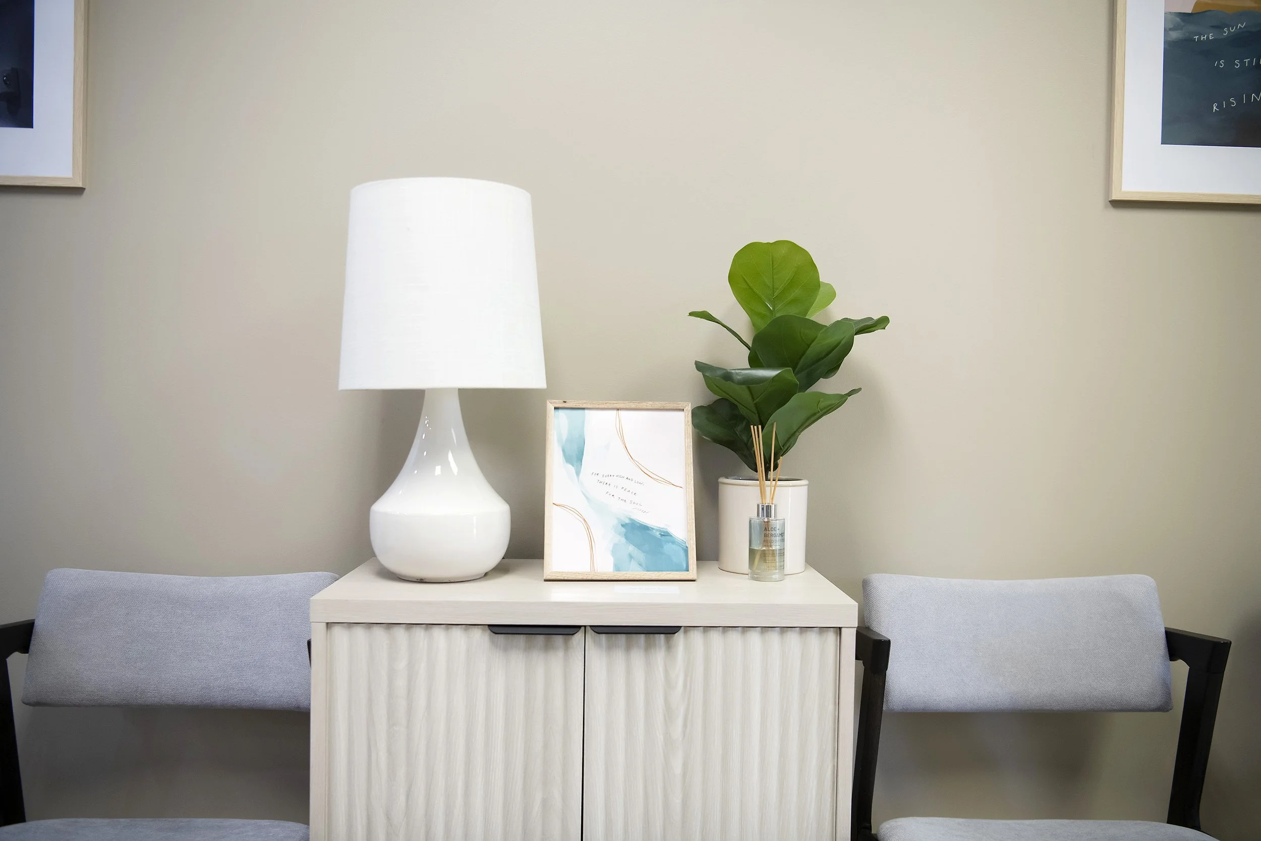 Desk with a white table lamp, a framed abstract art piece, a potted plant, and a reed diffuser between two gray chairs against a light-colored wall