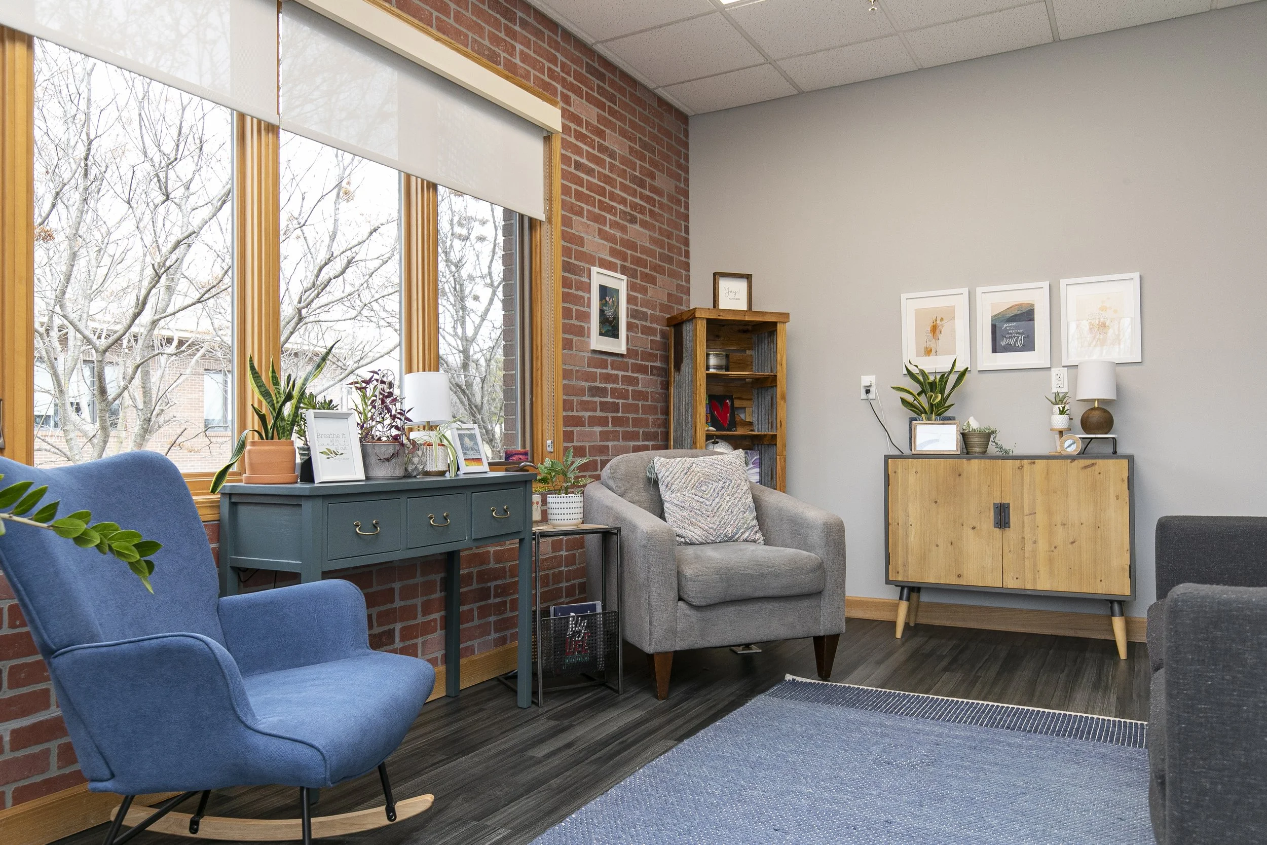 Therapy office with large windows, blue armchair, gray armchair with pillow, wooden cabinets with plants and decorative items, framed artwork on the wall, dark wood floor, and a blue rug.