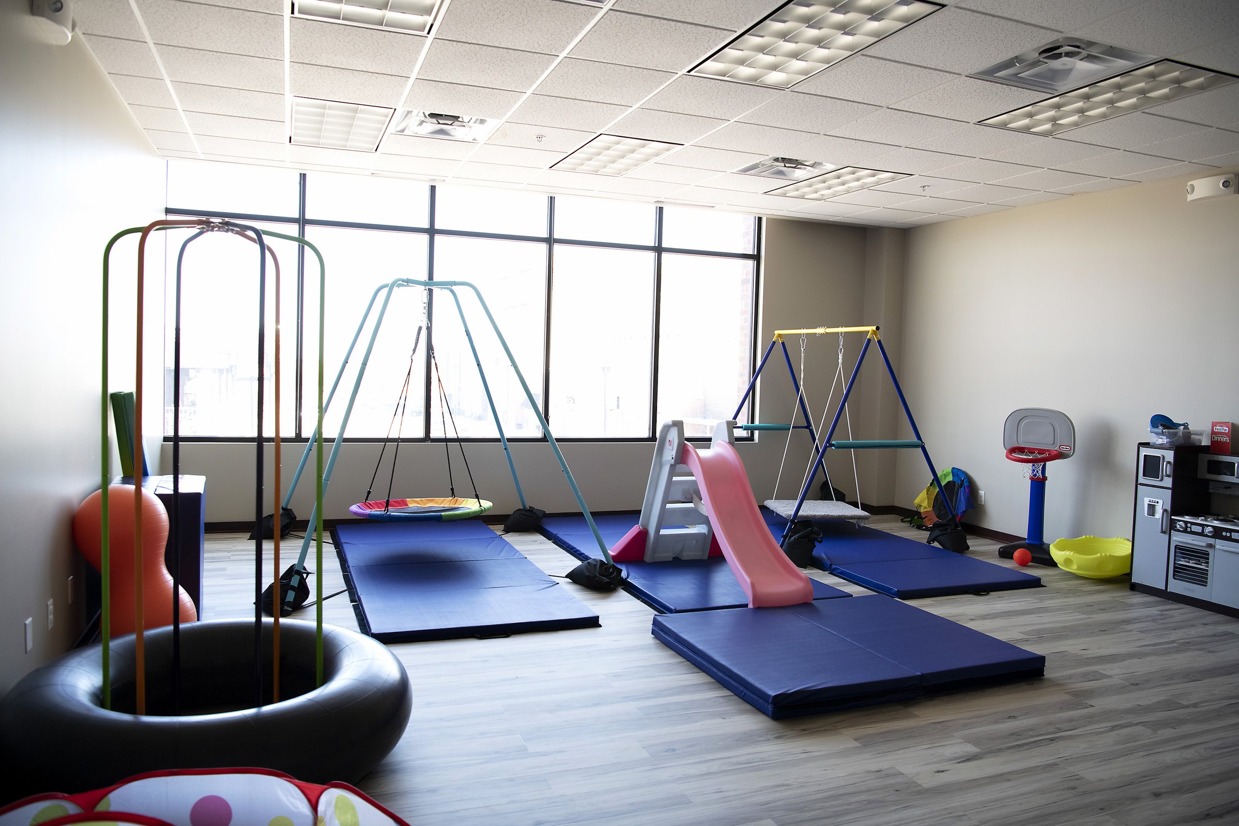 Indoor children's playroom with swings, slide, foam mats, basketball hoop, and toy kitchen.