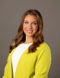 A woman with long brown hair, wearing a yellow blazer over a white top, smiling against a gray background.