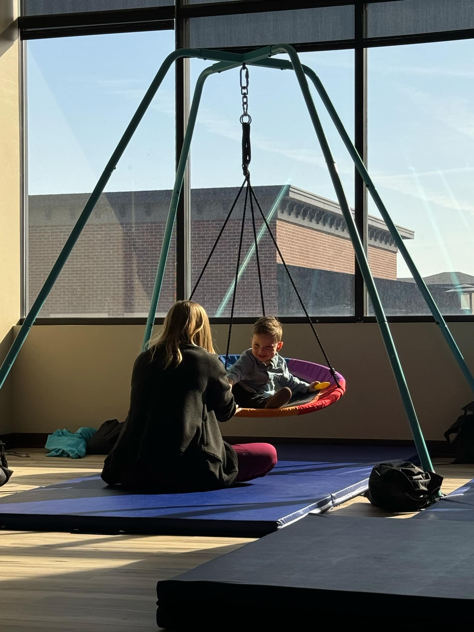 A woman and a young boy sitting on a blue mat inside a building next to a large window. The boy is sitting on a pink and purple swing, swinging gently indoors with a bright, sunny day visible outside.