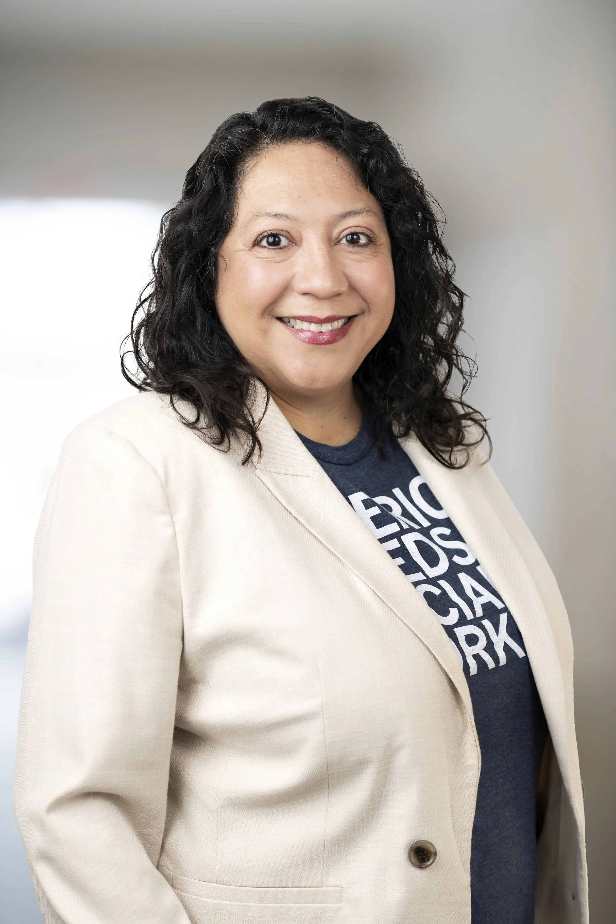 A woman with curly black hair smiling, wearing a beige blazer over a navy blue T-shirt with white text, standing indoors against a blurred background.