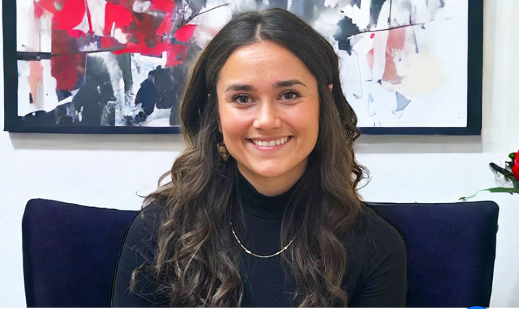 A young woman with long, wavy brown hair smiling at the camera, wearing a black turtleneck and a necklace, sitting in front of an abstract painting and a white wall.
