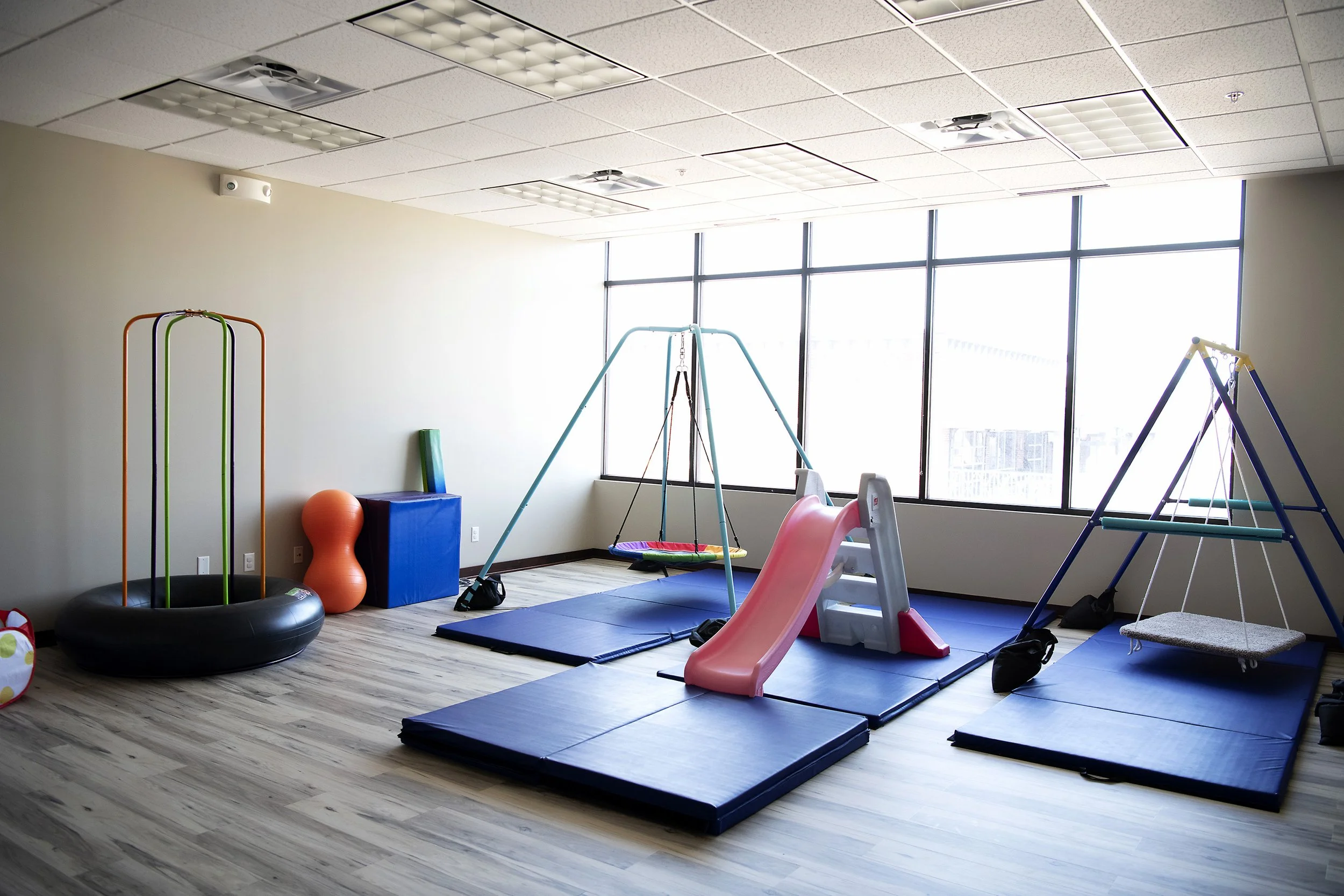 Children's indoor playroom with swings, slide, slide, foam mats, and soft play equipment near large windows.