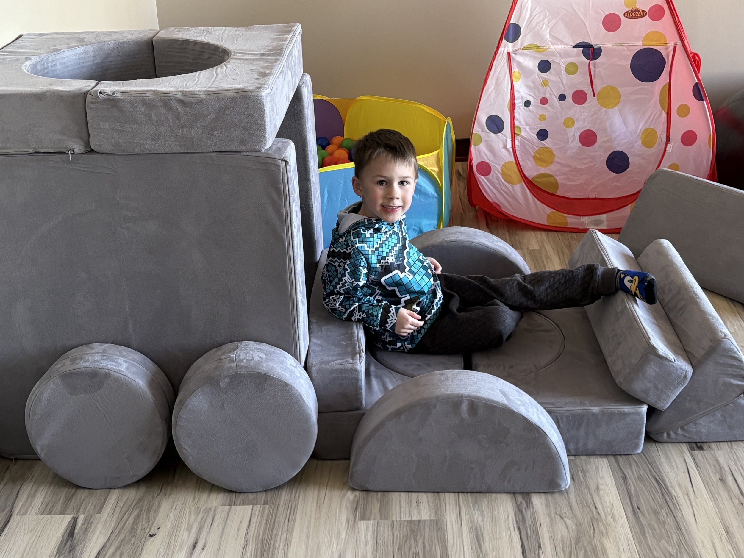 Young boy lying on a soft, gray toy train in a playroom, surrounded by colorful toys including a ball pit and a small fabric tent shaped like a tepee.