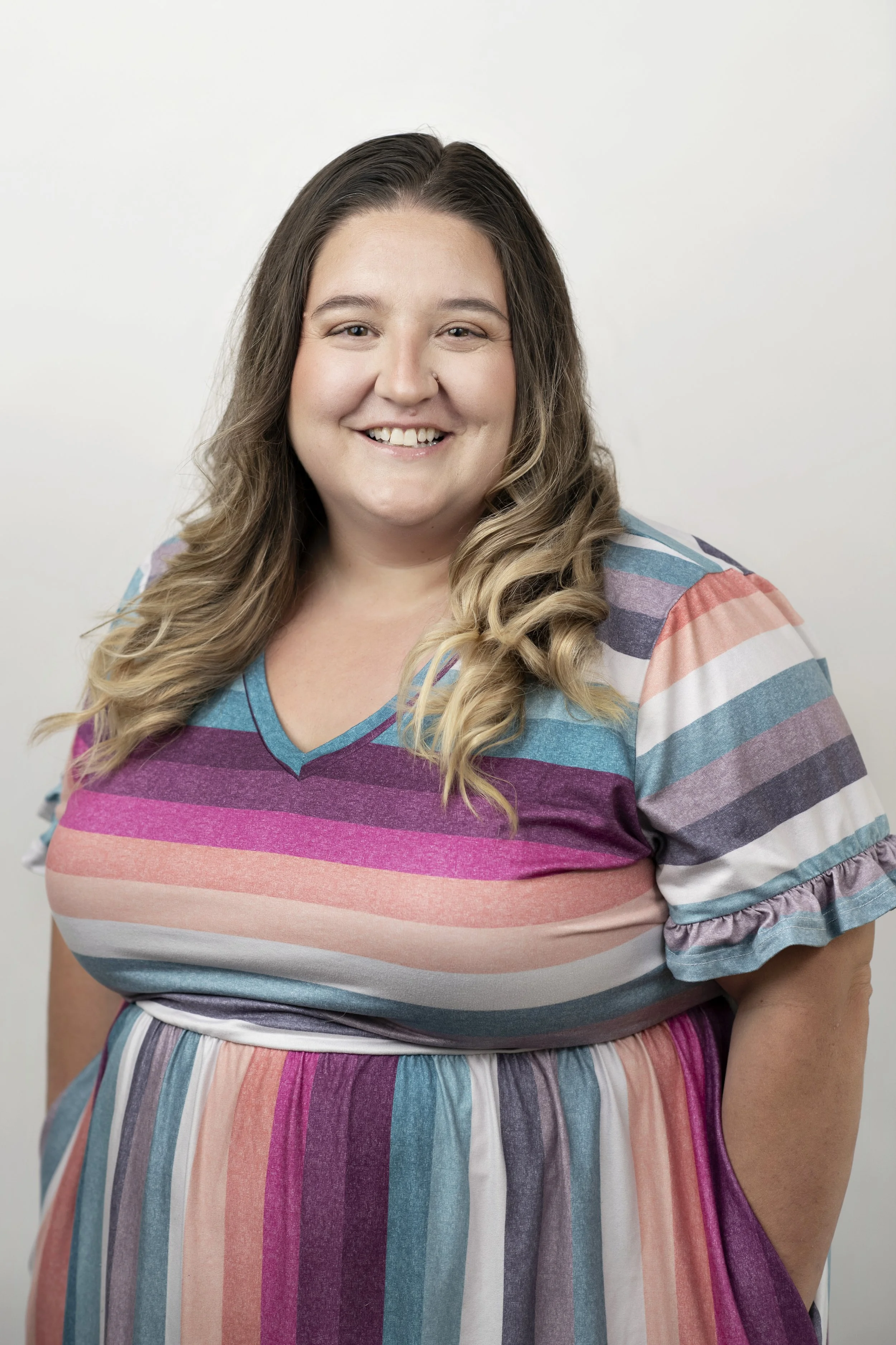 A smiling woman with long, wavy light brown hair wearing a colorful, striped dress with shades of purple, pink, blue, and white.
