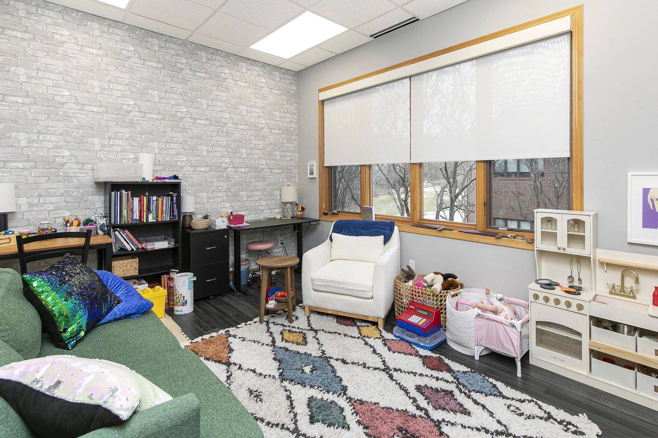 A cozy children's play therapy playroom with a green sofa, colorful pillows, a patterned rug, a white armchair, toy baskets, a toy kitchen set, and large windows with white blinds.