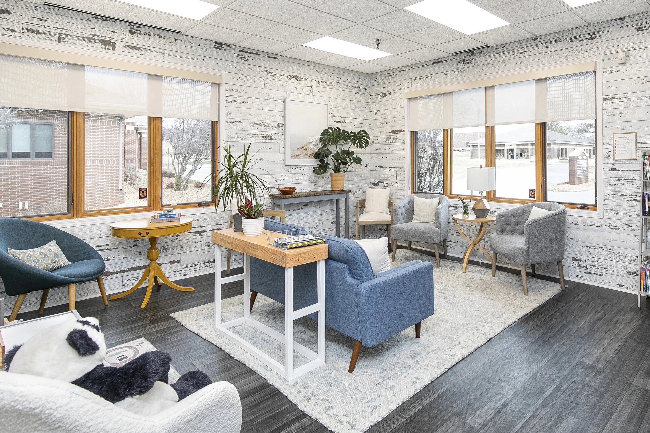 A cozy waiting room with whitewashed wooden walls, large windows, and various chairs and sofas, including a white plush dog on a white armchair.