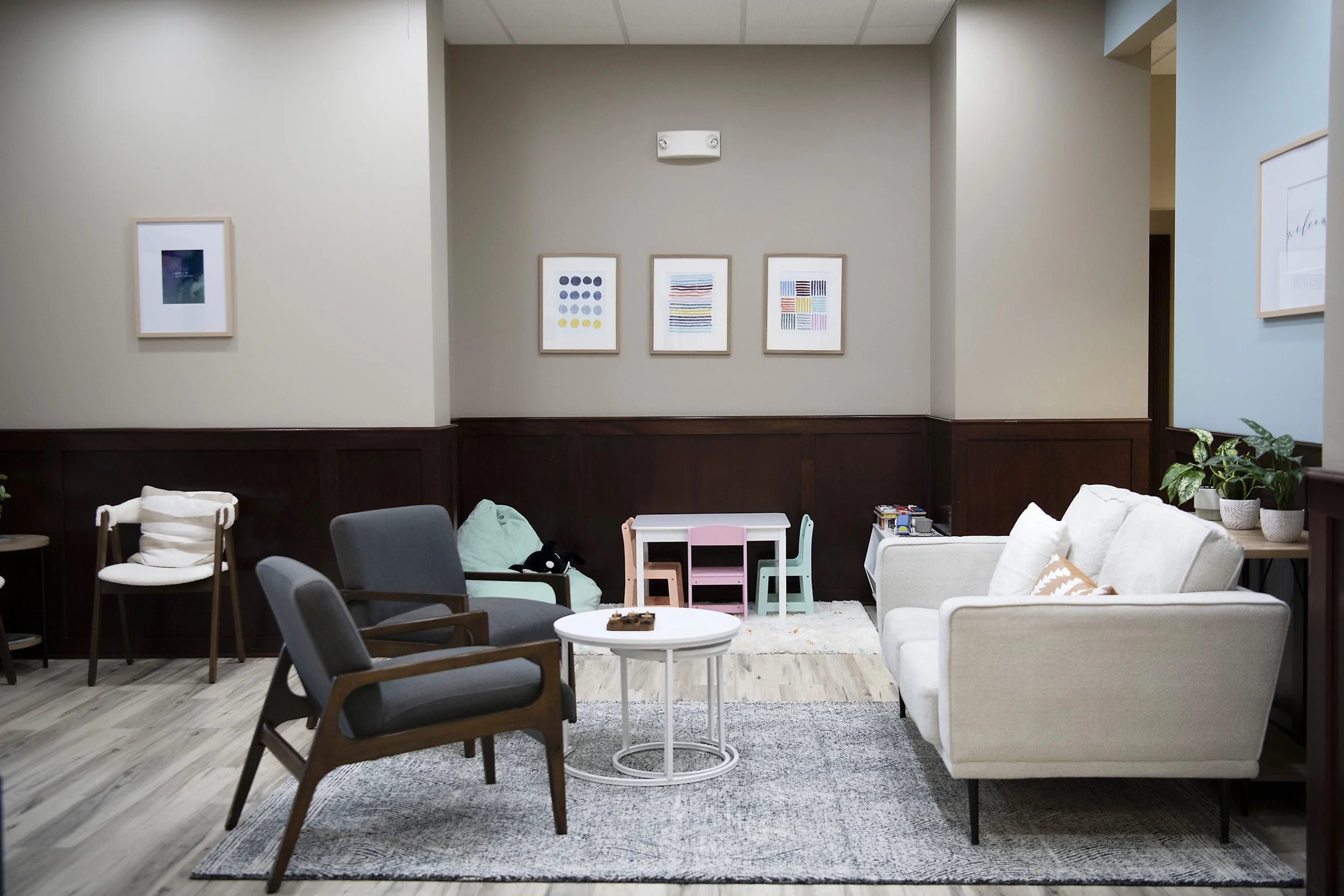 A cozy waiting area with various chairs, a white sofa, framed artwork on the walls, a small round coffee table, and a children's play table with chairs.