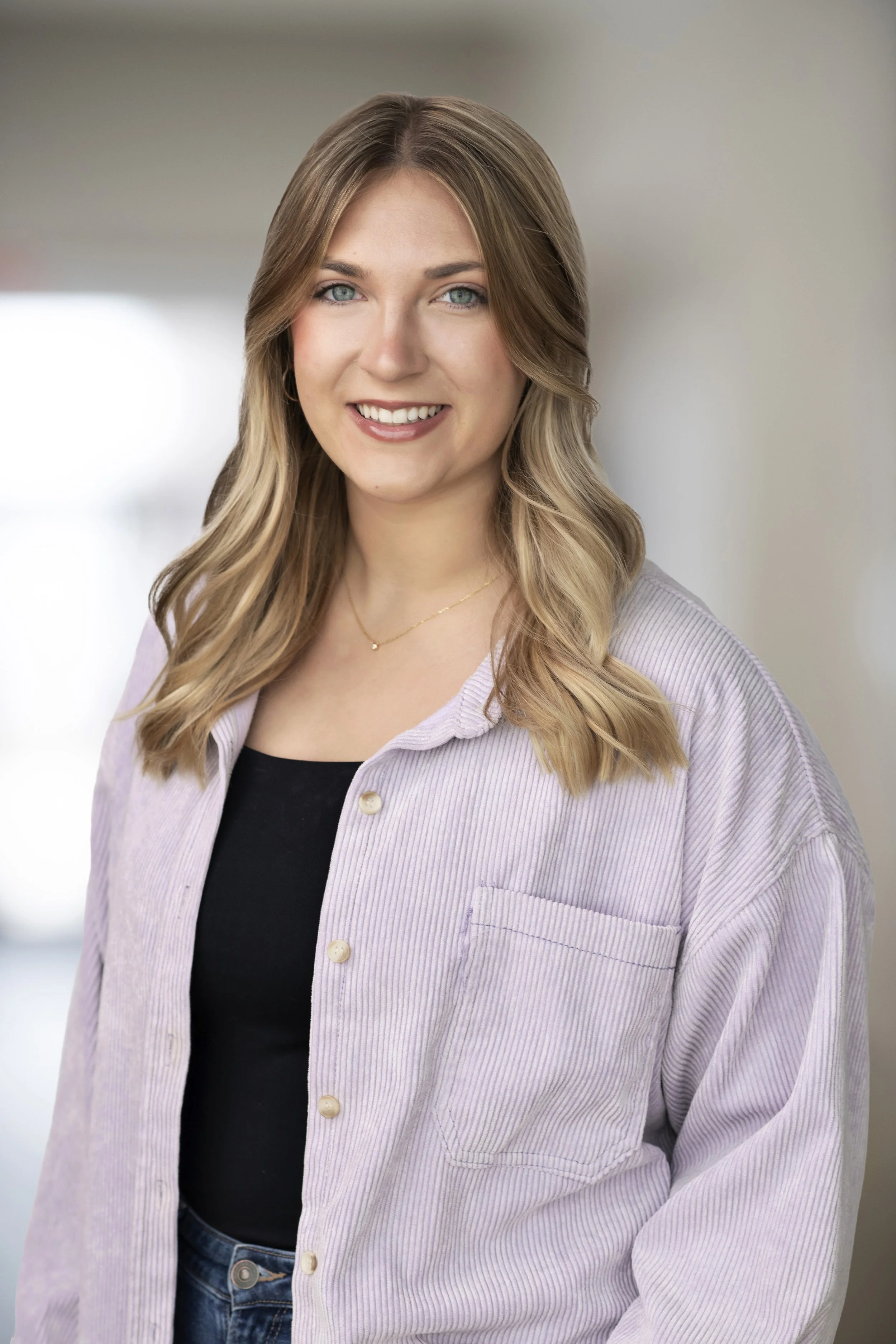 Young woman with blonde hair, blue eyes, wearing a black top and a light purple striped shirt, smiling in an indoor setting.