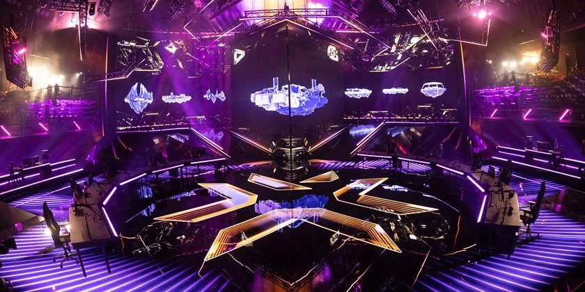 A stage with bright purple and pink neon lights, large screens showing space and spaceship graphics, and multiple workstations, set up for a live event or concert.