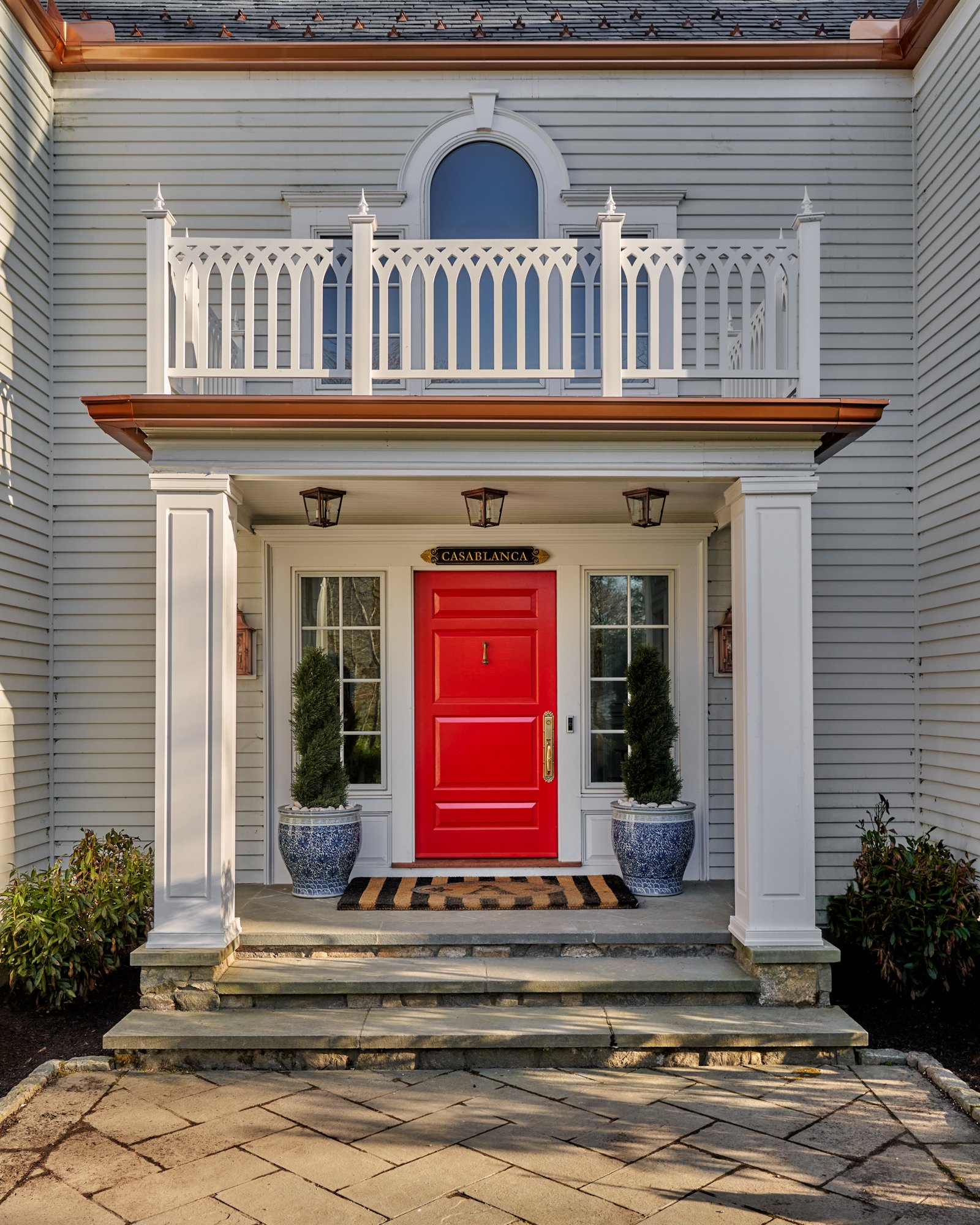 A bold red lacquered door sets the tone for this stately yet spirited entryway, framed by classic white millwork, gas lanterns, and fluted columns. Above, a Gothic-inspired balustrade adds architectural flourish, while chinoiserie planters and a geom