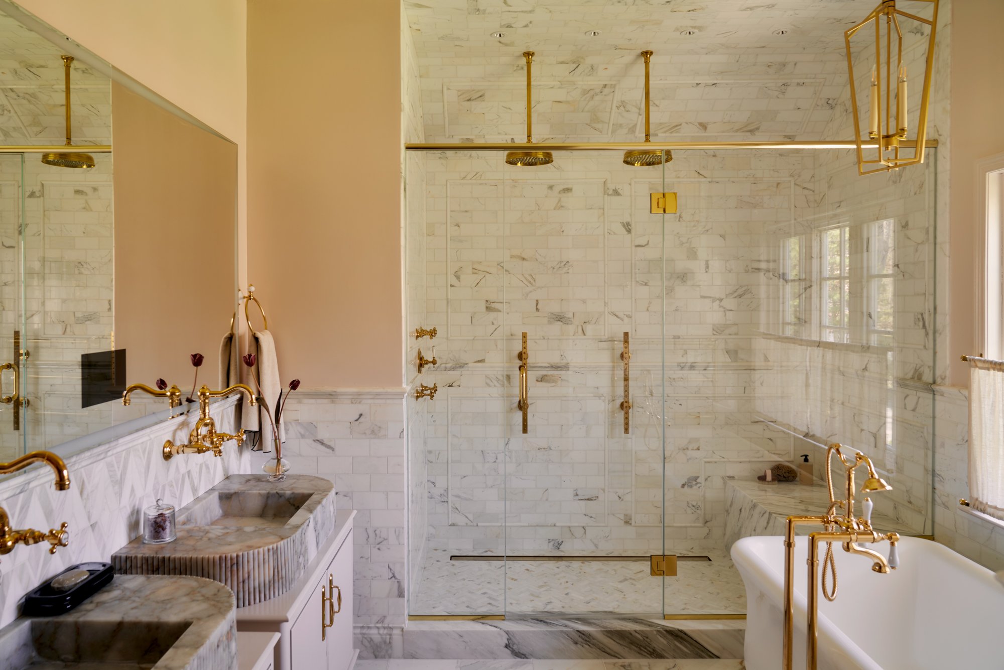A harmonious blend of soft blush tones, honed stone, and gleaming brass defines this serene primary bath. Fluted marble vanities sit beneath a full-length mirror, their sculptural forms echoing the geometry of the chevron tile backsplash. Dual rainfa