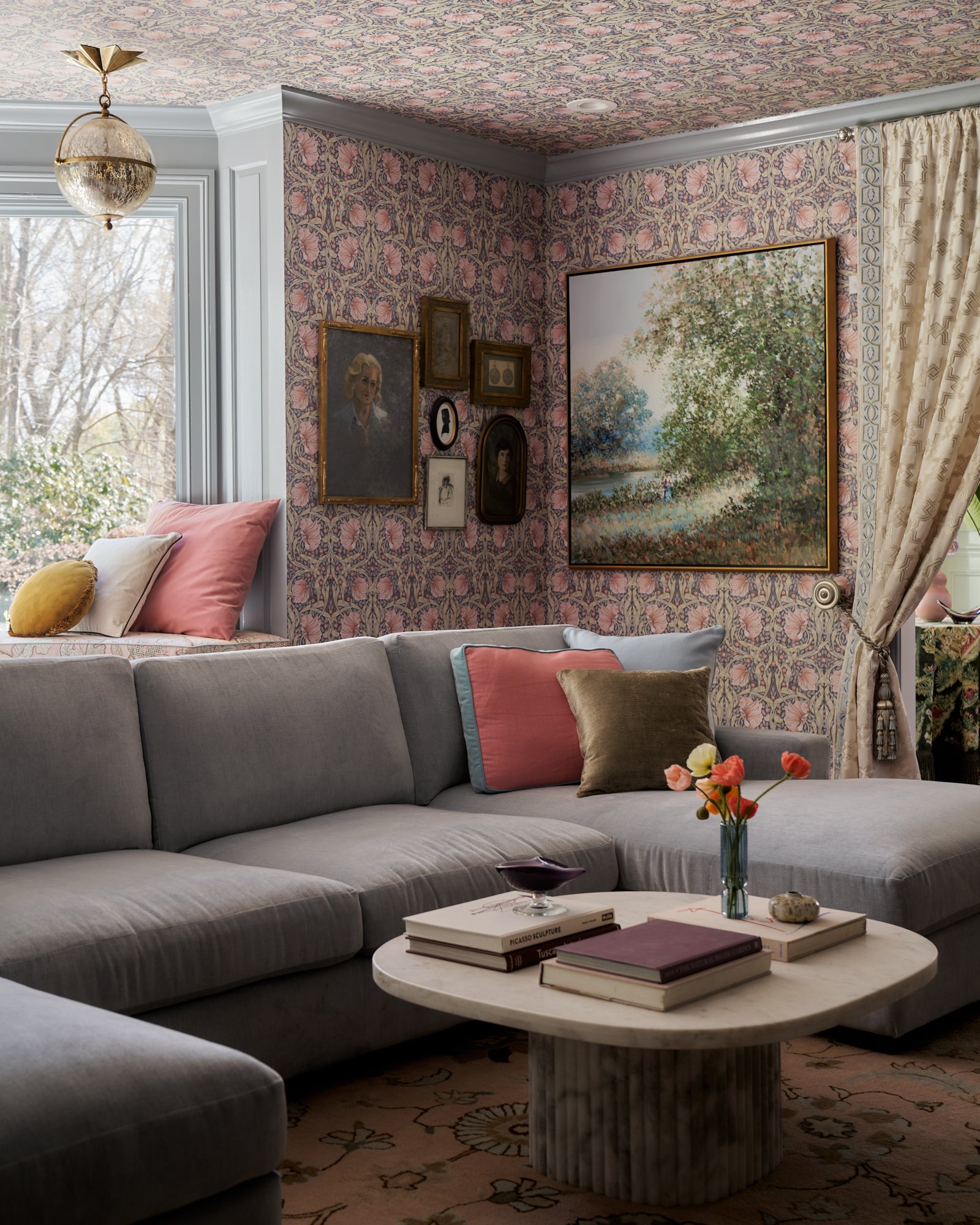 With walls and ceiling swathed in a rose-strewn William Morris print, this parlor blurs the boundary between architecture and ornament. A modern linen sectional grounds the space with generous scale, while vintage silhouettes, pastoral landscapes, an