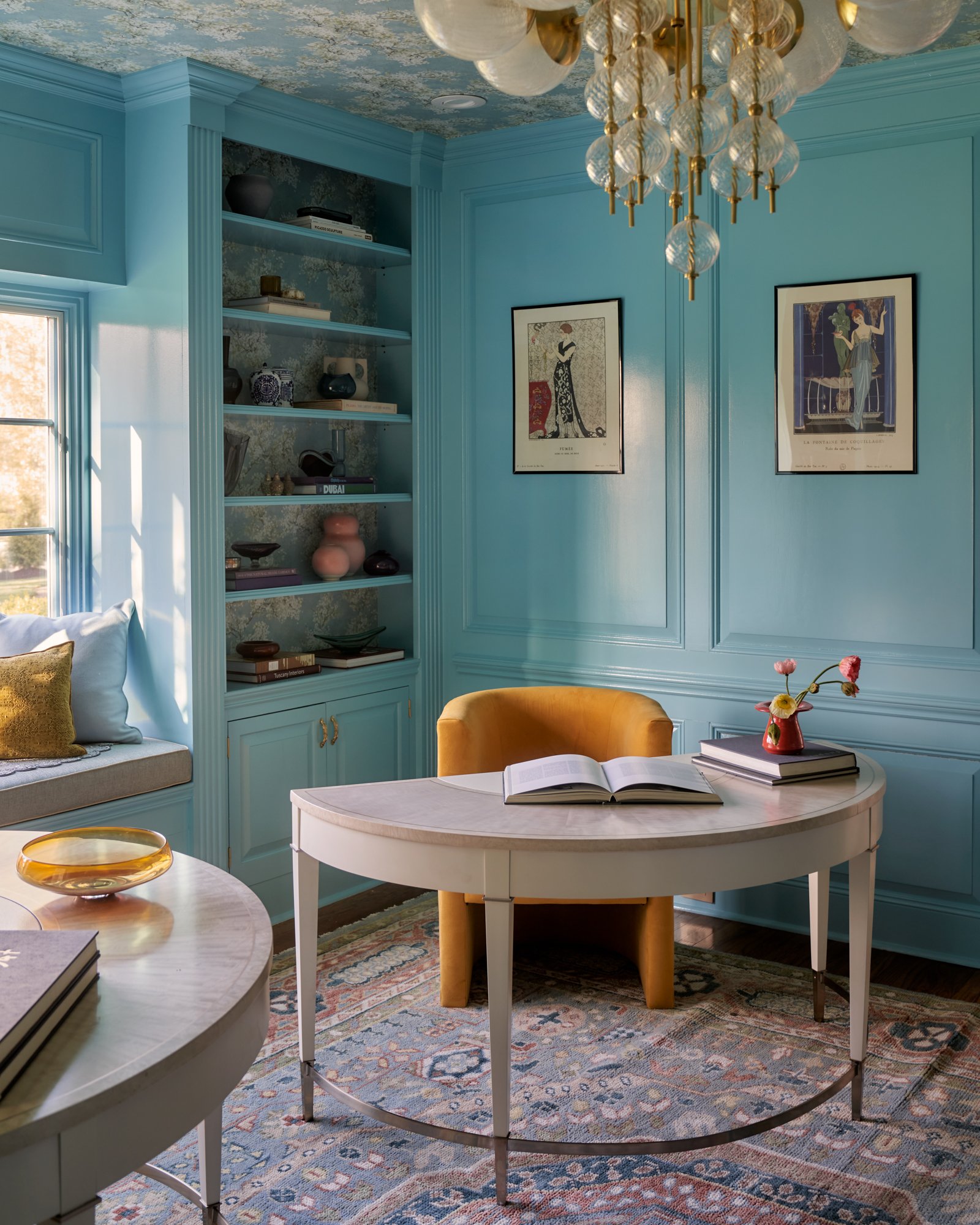 This intimate view of the cerulean study reveals the layered beauty of its details—from the demi-lune desks topped in creamy stone to the ochre velvet tub chair offering both comfort and contrast. Built-in shelving is backed with subtle floral wallpa