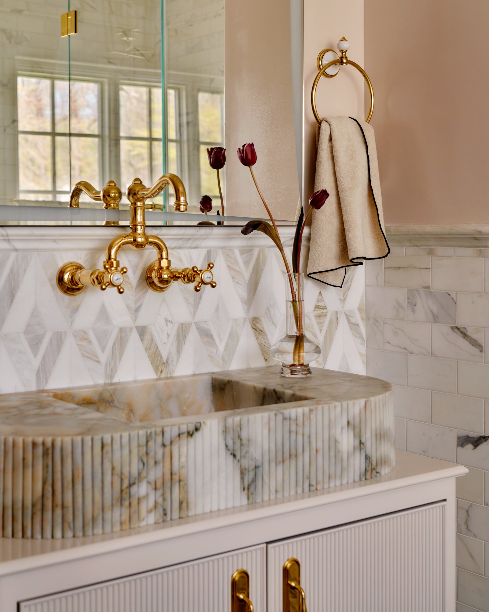 A custom fluted marble vanity commands attention with its sculptural presence, complemented by a faceted mosaic backsplash in honed stone. Warm blush walls and unlacquered brass fixtures add depth and elegance, while a single vase of dark tulips offe
