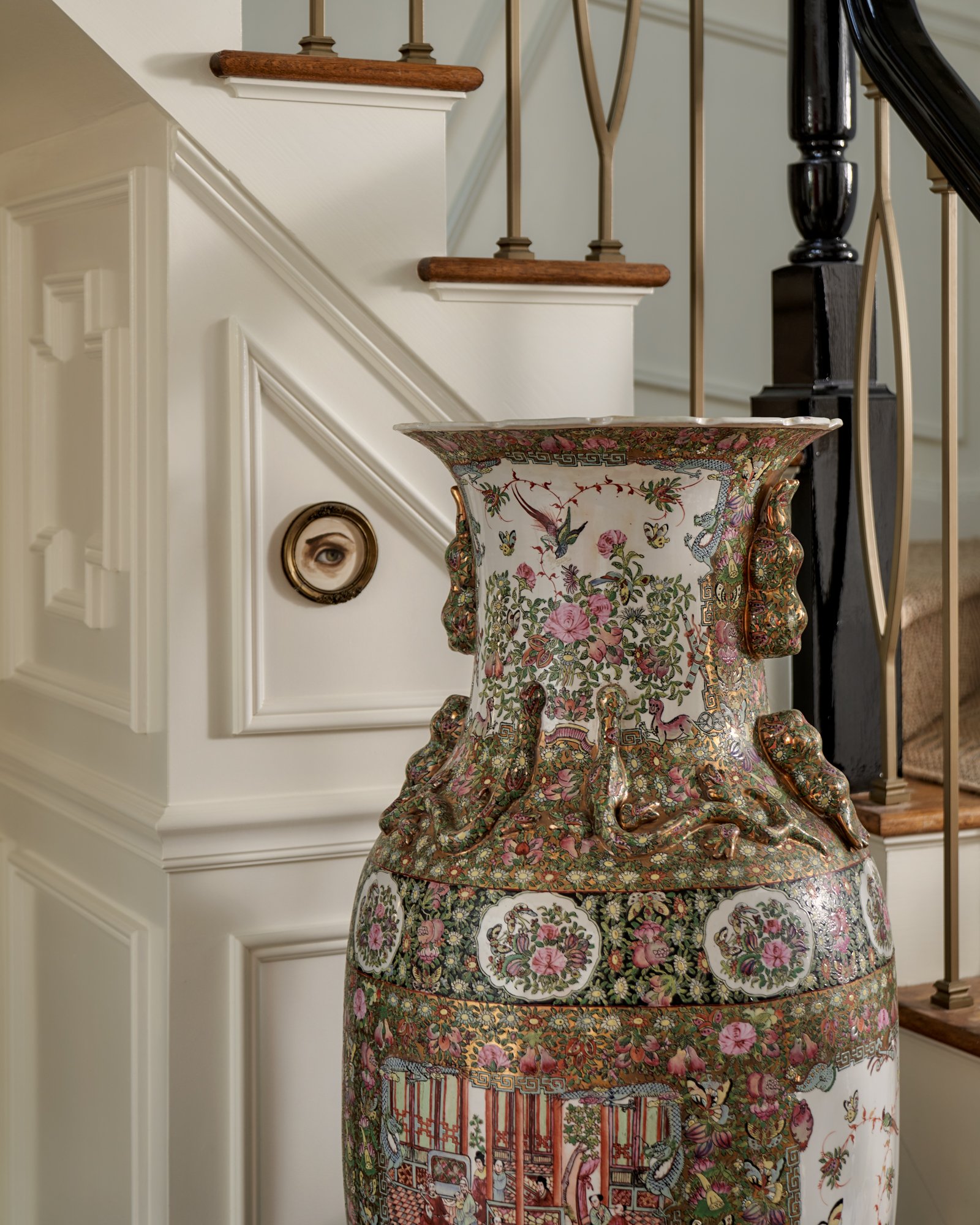 A towering Famille Rose vase commands attention at the stair’s base, its intricate narrative and gilded florals set against crisp ivory millwork. Above it, a single antique eye miniature—piercing and poetic—adds a surrealist wink to the traditional s