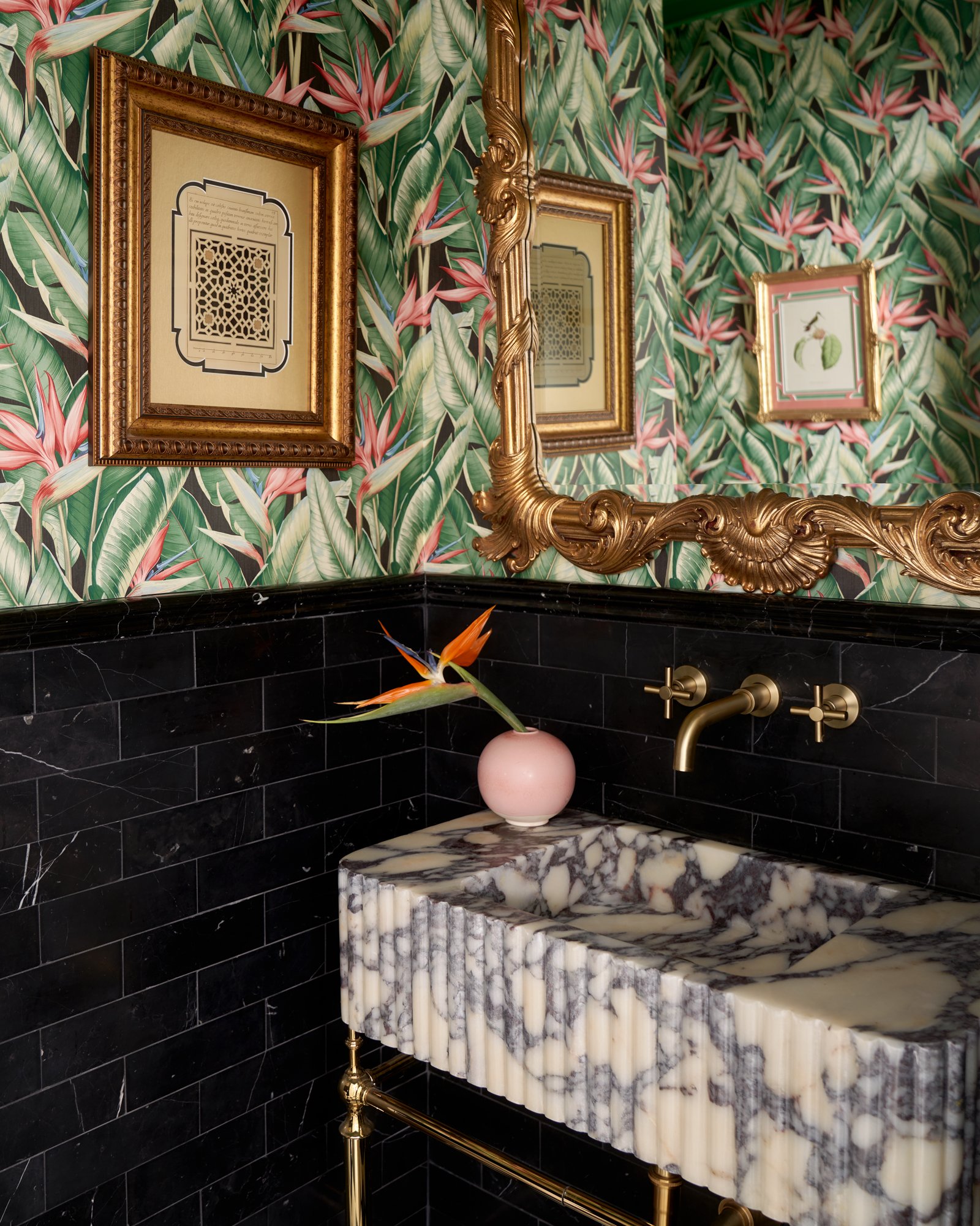 This jewel-box powder room marries bold pattern with sculptural sophistication. Jungle-inspired wallpaper climbs the walls, wrapped in ornate gilt framing and crowned by a carved Rococo mirror. A dramatically veined, fluted marble sink floats above a