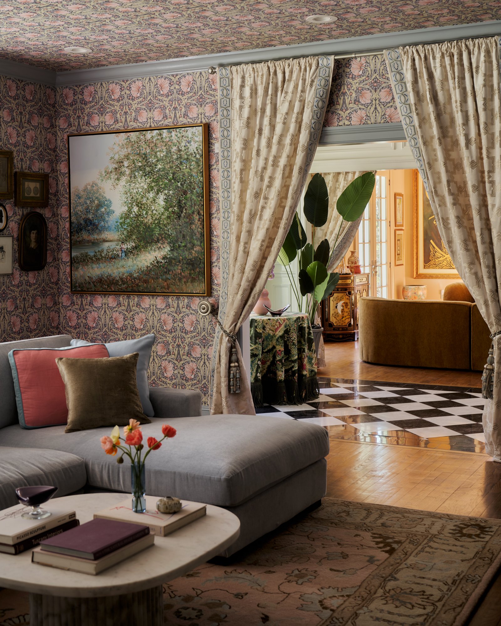 A romantic parlor swathed in floral wallpaper opens through tasseled silk drapery into a luminous salon of palms, lacquer, and gold. The juxtaposition of soft pastels and graphic black-and-white stone flooring creates a cinematic moment of contrast a
