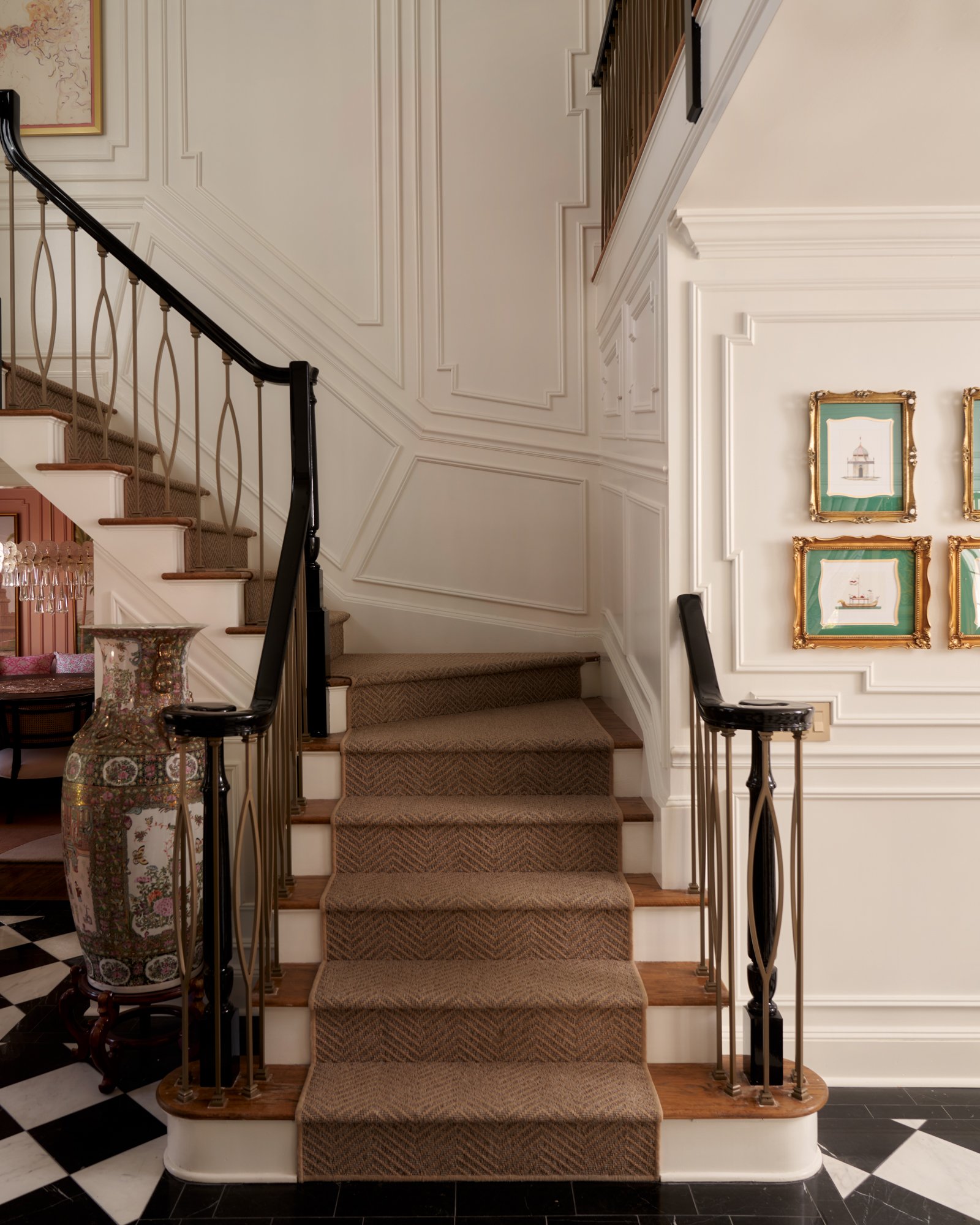 This sculptural stair hall is a study in contrast and quiet detail. Crisp panel molding climbs the walls in a rhythmic dialogue with the iron-and-brass balustrade, while a tailored wool stair runner in a subtle chevron pattern softens the ascent. The