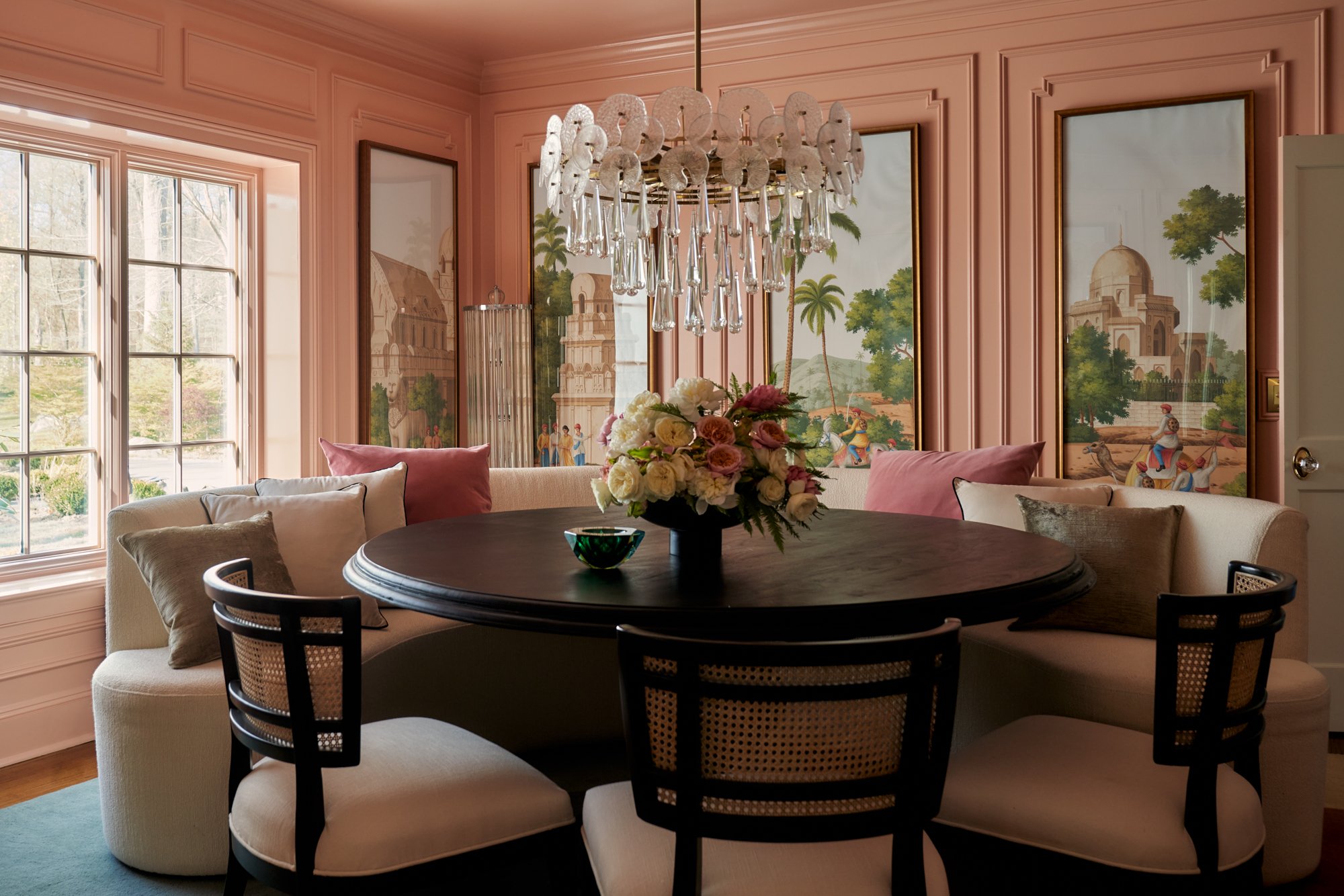 An ode to romance and storytelling, this dining room is enveloped in blush paneling and accented with hand-painted scenic panels evoking the architecture of empire and the lushness of faraway lands. A tailored curved banquette wraps a matte black ped