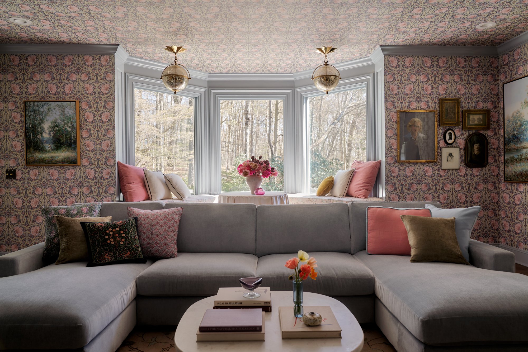 This moody-yet-whimsical parlor envelops you in pattern and nostalgia, with a rose-and-vine wallpaper extended across both walls and ceiling in a fully immersive gesture. A pale lavender sectional anchors the room with softness, flanked by jewel-tone