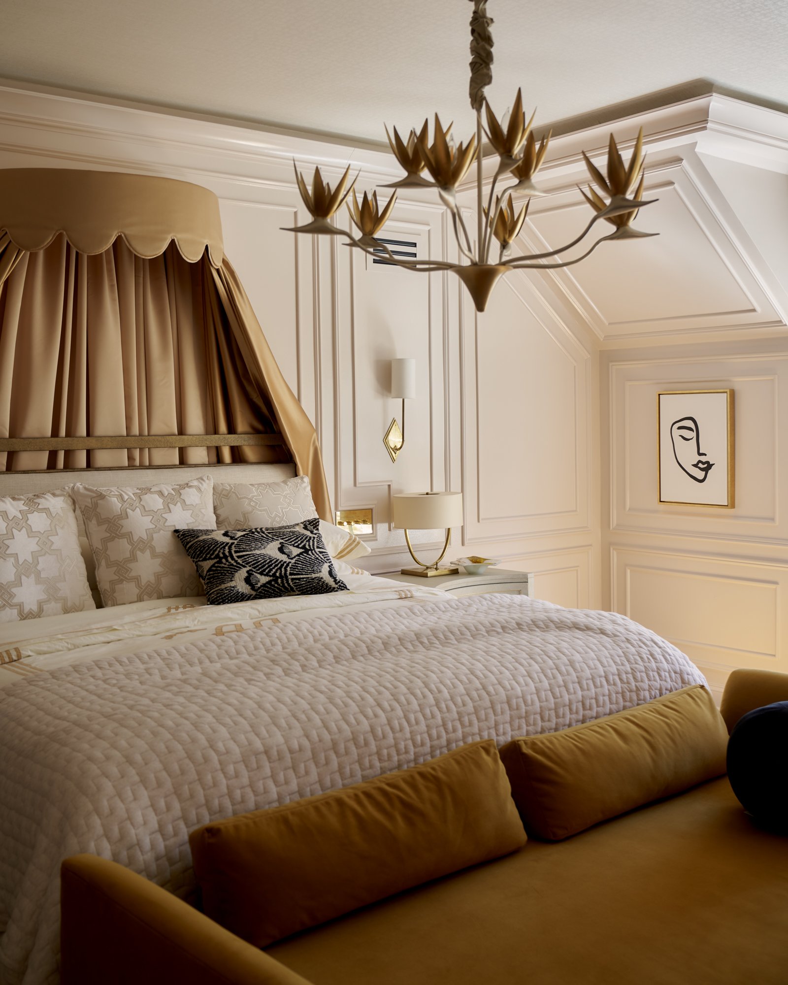 Layered with softness and structure, this primary bedroom pairs architectural millwork with a scalloped canopy and a sculptural chandelier that evokes blooming florals in flight. A palette of ivory, camel, and burnished gold wraps the room in warmth,