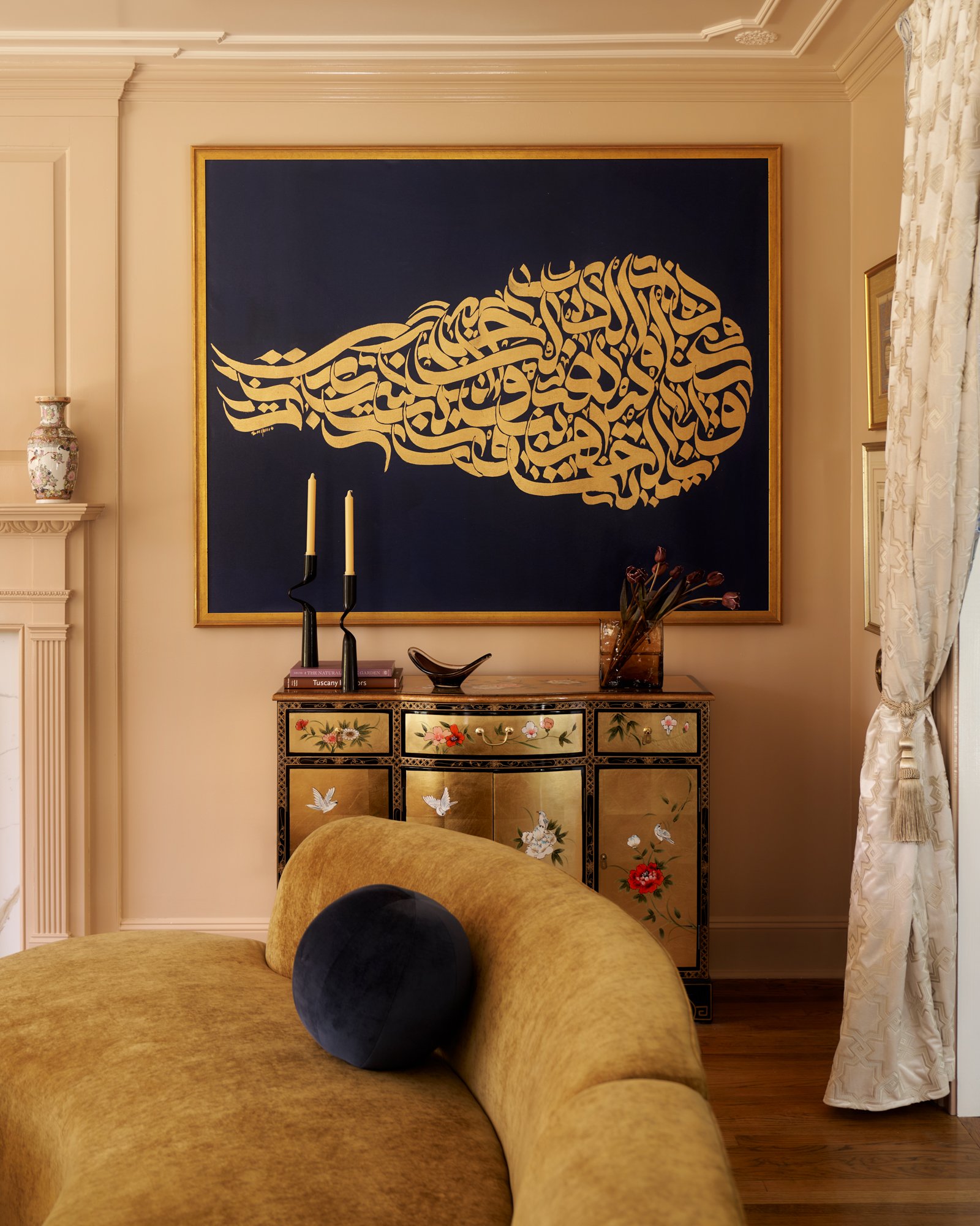 A moment of high contrast and cultural layering—this vignette pairs a sweeping navy-and-gold calligraphy canvas with a hand-painted chinoiserie chest, creating a dialogue between East and Middle Eastern artistry. The curved ochre velvet sofa echoes t
