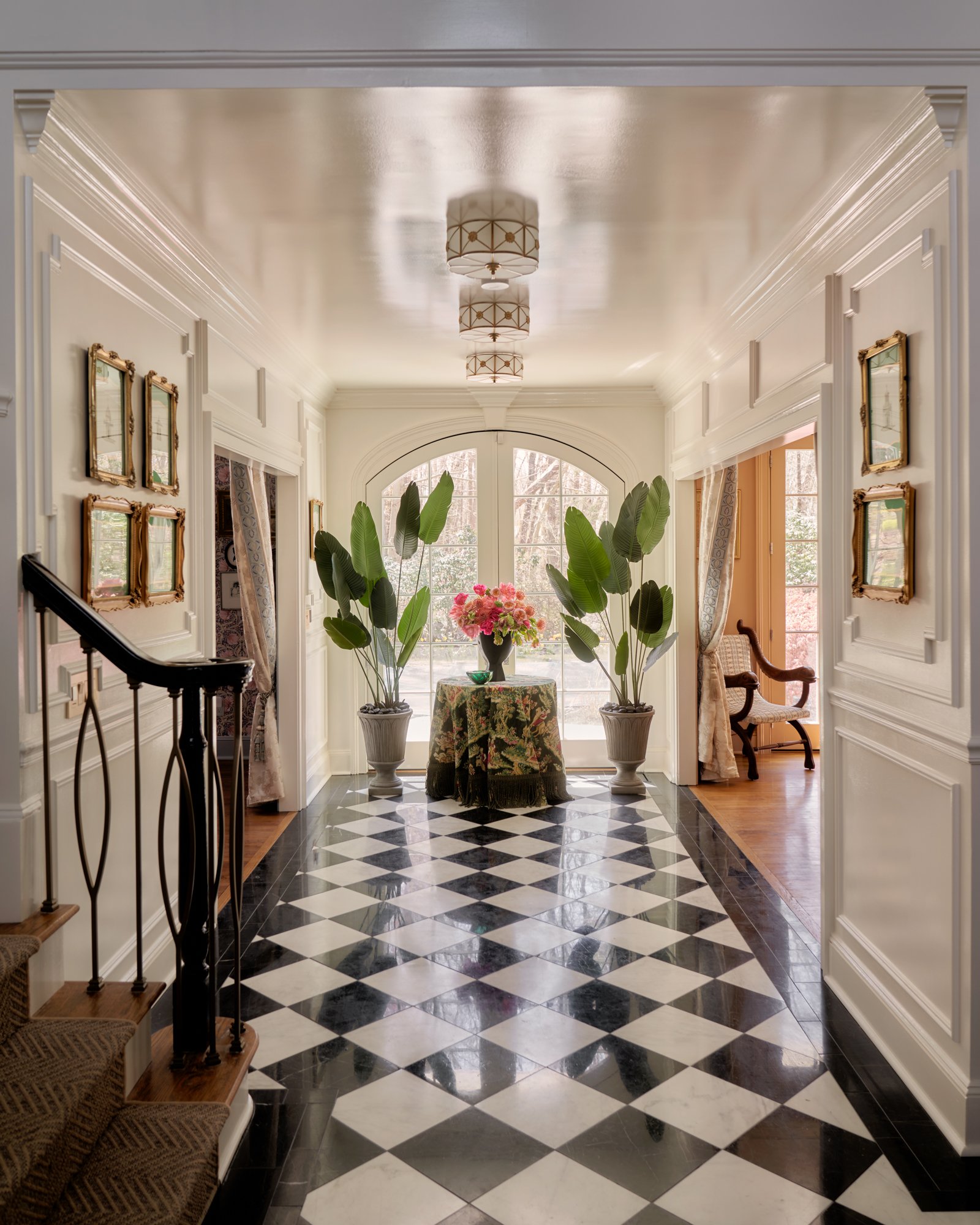 This statement entryway sets the tone for the home’s layered aesthetic—a study in symmetry, glamour, and botanical drama. Classic black-and-white checkerboard marble stretches the length of the hall, drawing the eye toward arched French doors and a l